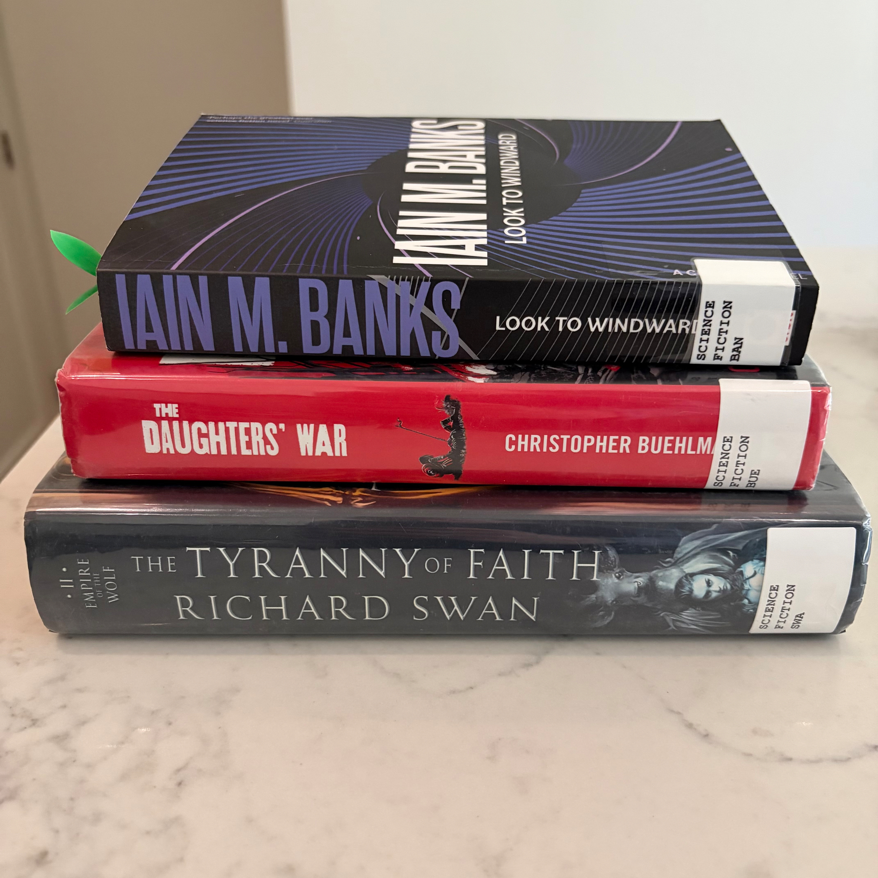 A stack of three books titled Look to Windward by Iain M. Banks, The Daughters' War by Christopher Buehlman, and The Tyranny of Faith by Richard Swan is placed on a marble surface.