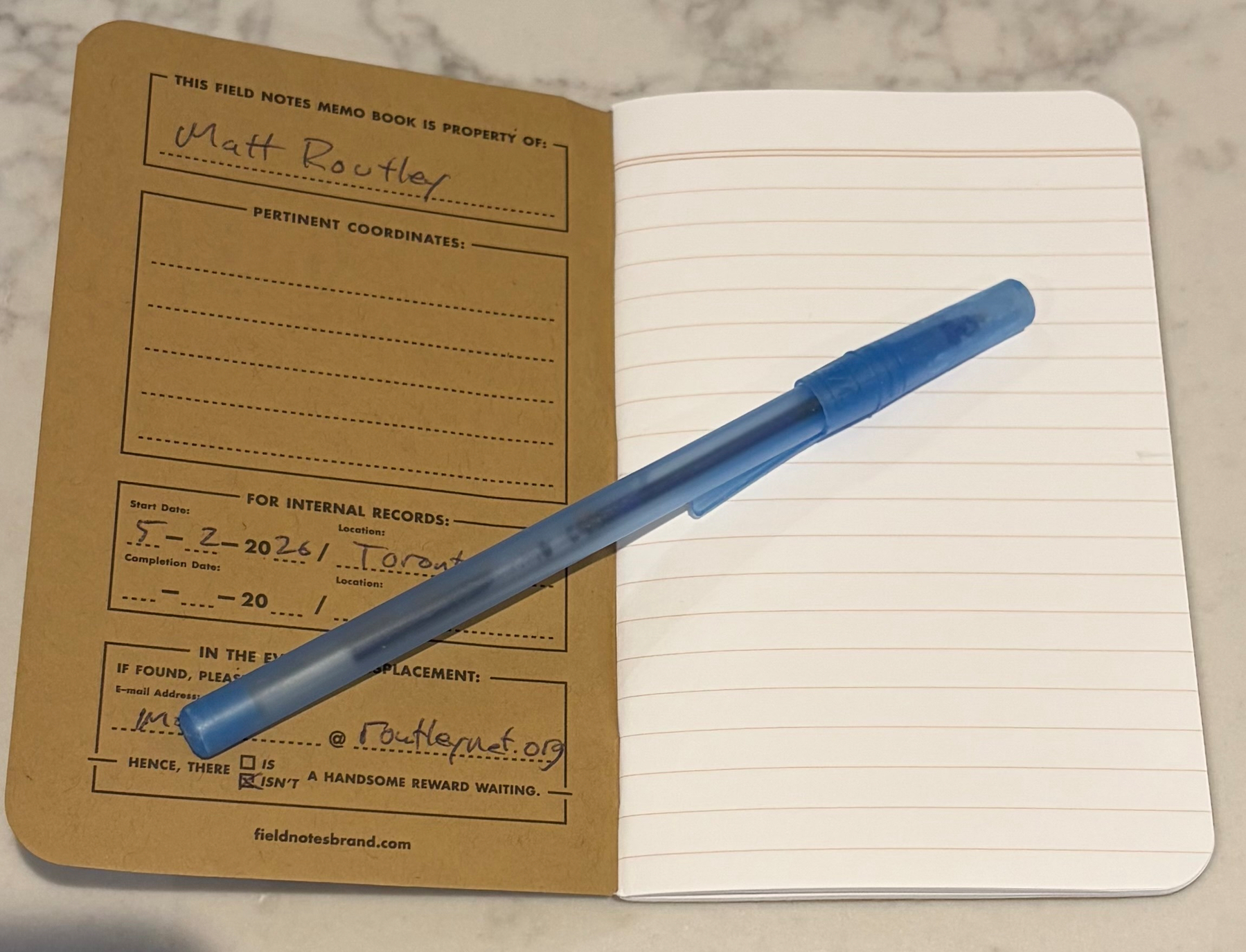 An open Field Notes memo book, starting on a blank first page, is accompanied by a blue pen resting on its pages.