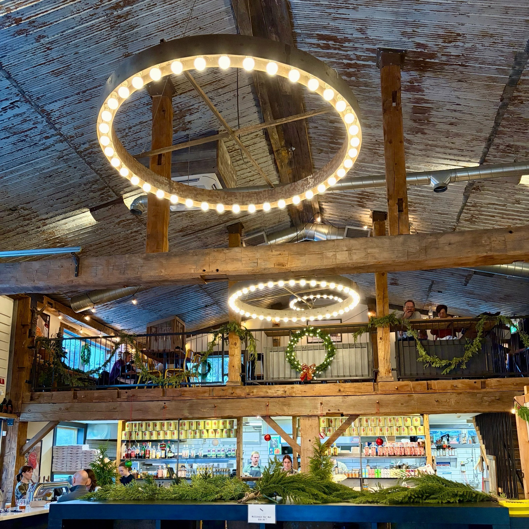 A multi-level restaurant or café interior with rustic industrial design featuring exposed wooden beams, corrugated metal ceiling panels, and two large circular LED light fixtures hanging from the ceiling. The space includes an upper mezzanine level with wooden railings and seating areas, decorative greenery garlands, and a service counter area below with plants and holiday decorations. The warm lighting and natural wood create an inviting atmosphere.​​​​​​​​​​​​​​​​