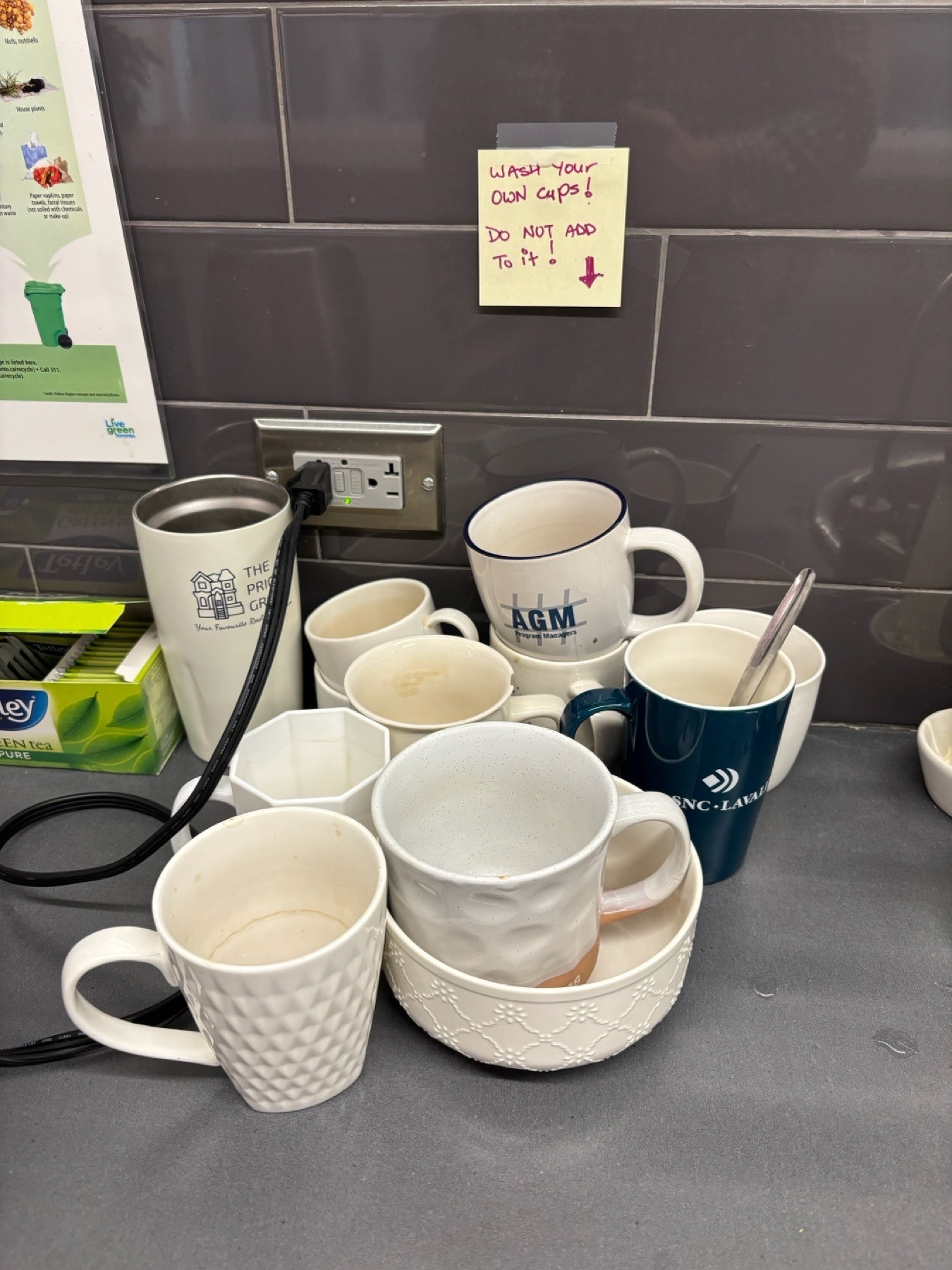 A pile of dirty cups, beside a sink, with a sticky note saying “Wash your own cups! Do not add to it!”