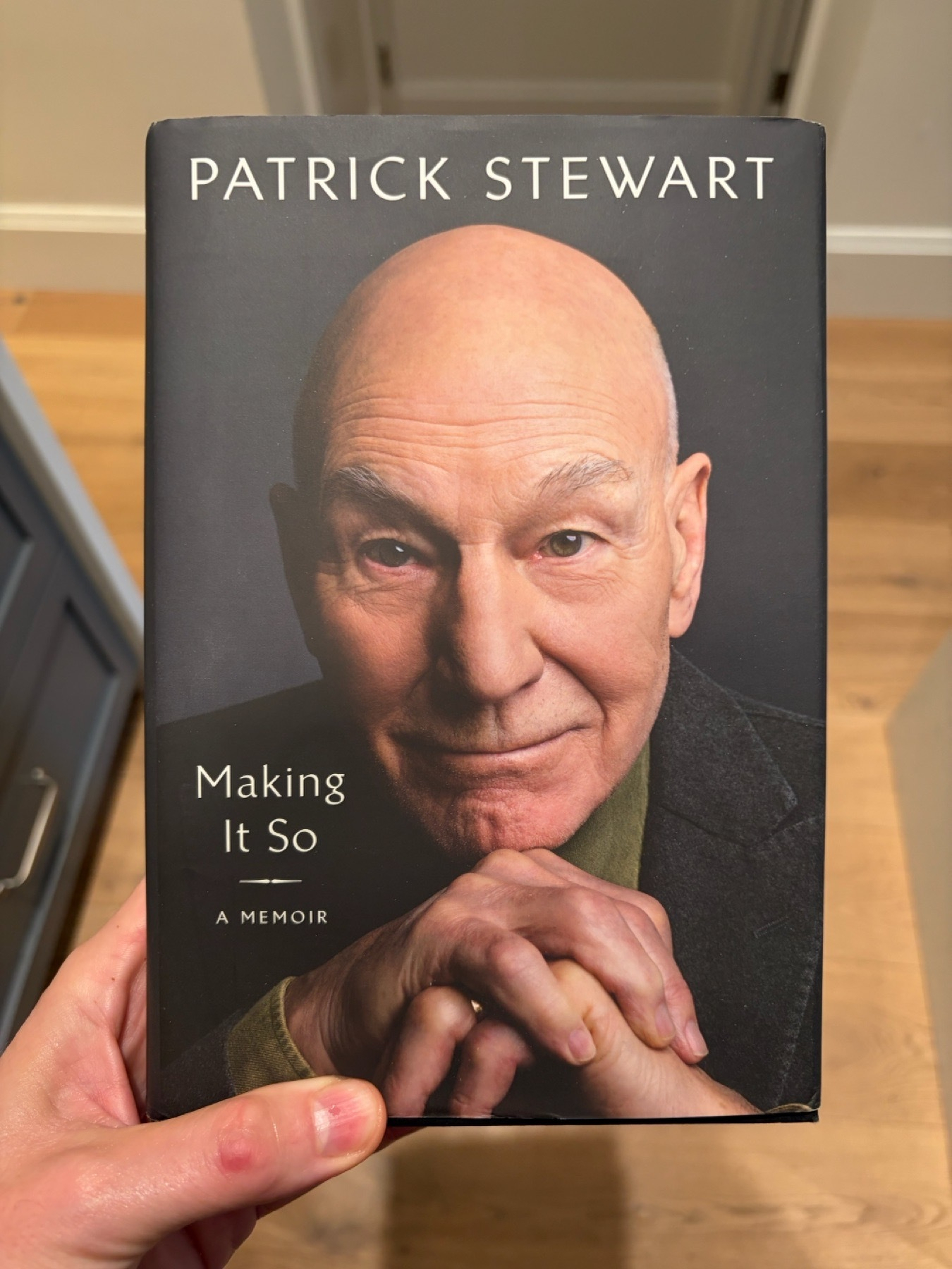 A person holds a memoir titled &ldquo;Making It So&rdquo; by Patrick Stewart. The cover features a portrait of the author wearing a dark jacket and a green sweater, with the title and author&rsquo;s name printed at the top and left side.