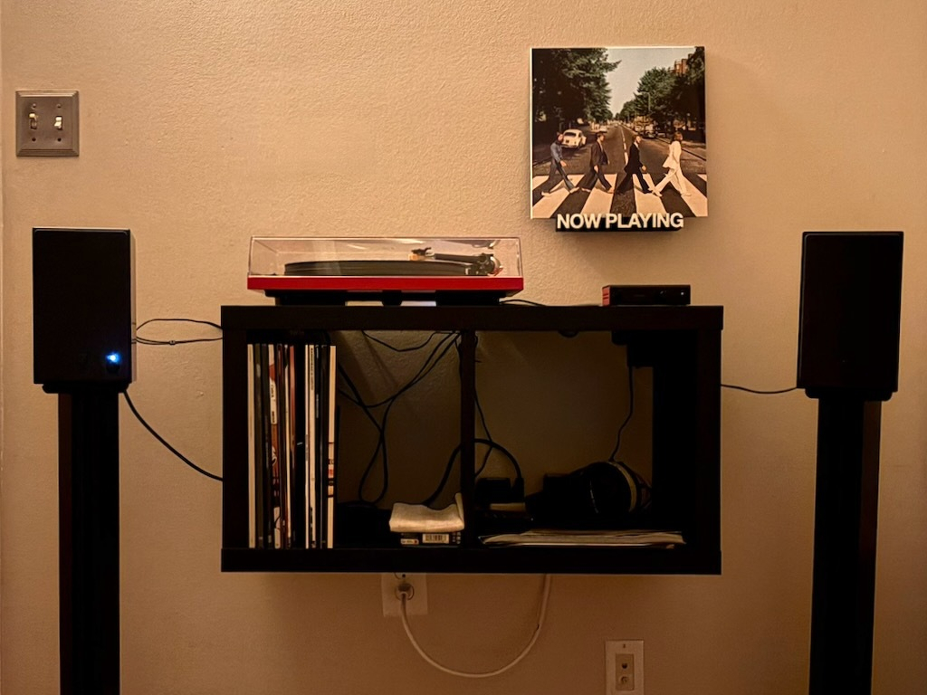 A turntable setup with speakers and vinyl records is neatly arranged on a mounted shelf, featuring the Abbey Road album cover with the text NOW PLAYING underneath.