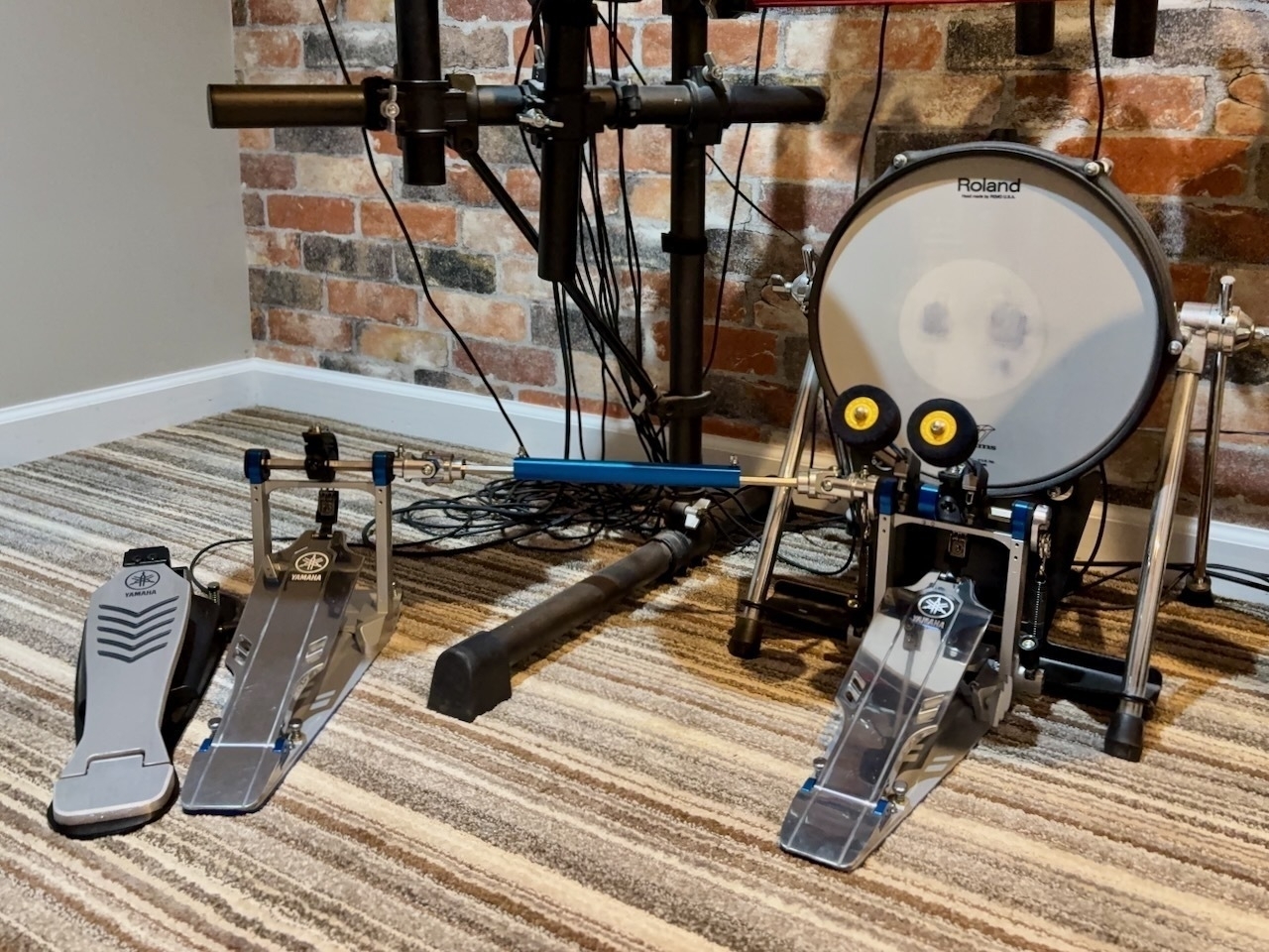 Yamaha drum pedals and a Roland V-Drum kick pad drum are positioned on a carpeted floor in front of a wall with brick wallpaper design.