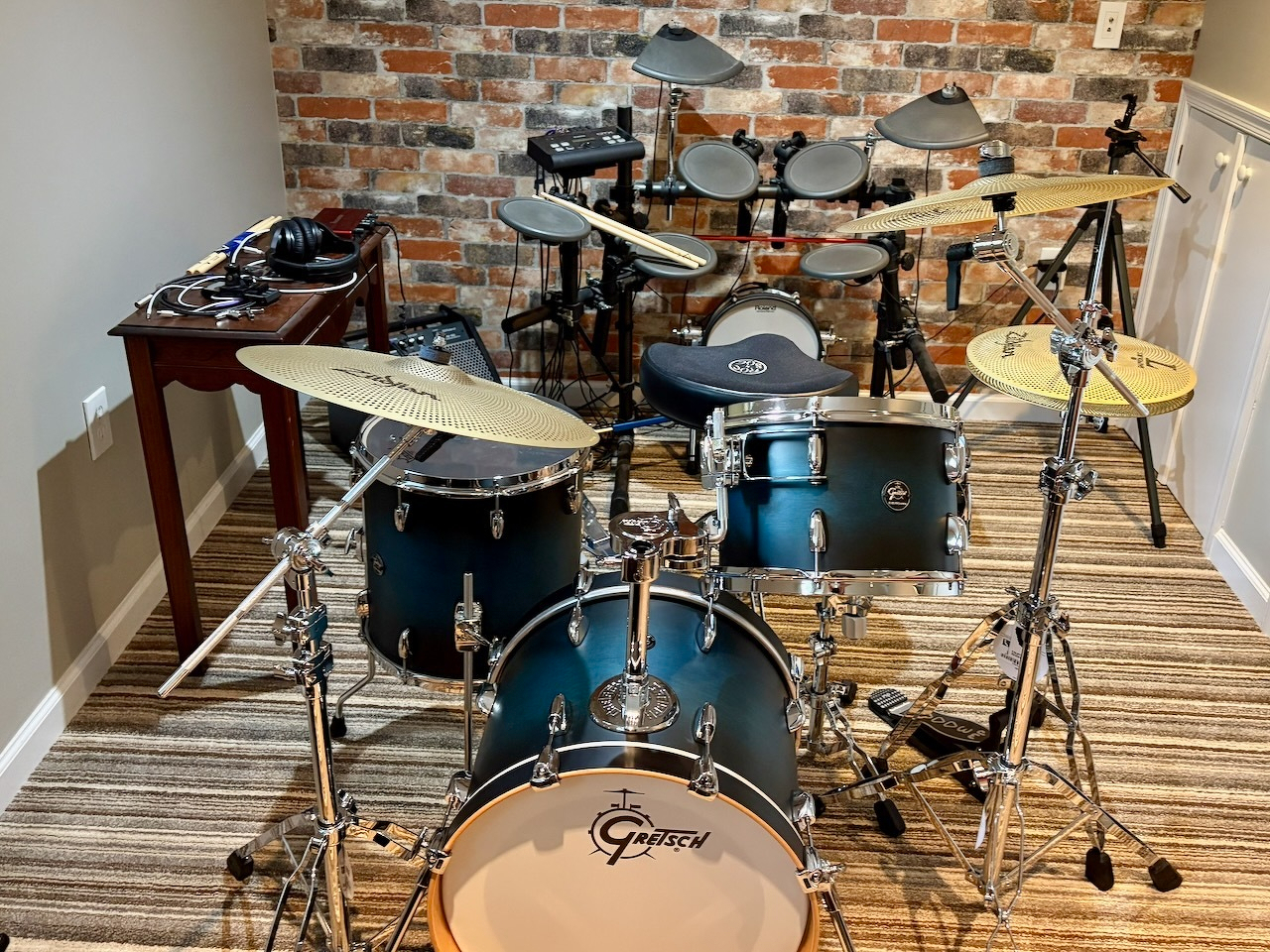 A Gretsch 4-piece acoustic drum kit and a Yamaha 5-piece electronic drum set are set up in a room with brick-patterned wallpaper.