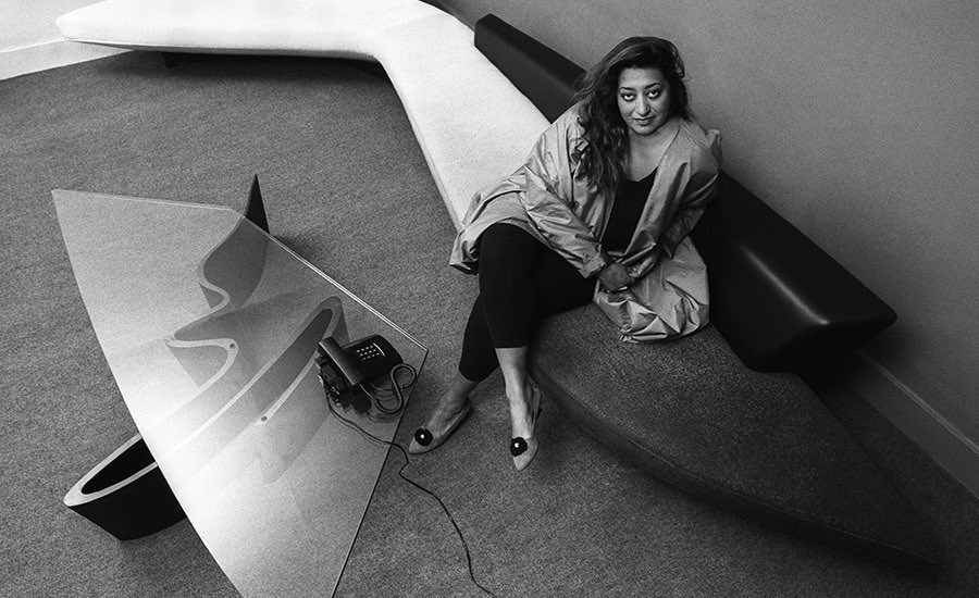 Black and white photo of Zaha Hadid sitting on a bench next to a glass coffee table. There is a corded phone on the table.