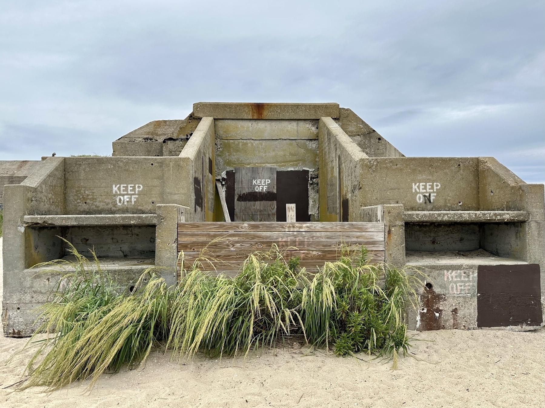 Photo of a concrete bunker on a sandy beach, grass growing around it, with “Keep off” graffiti on 3 of the walls.