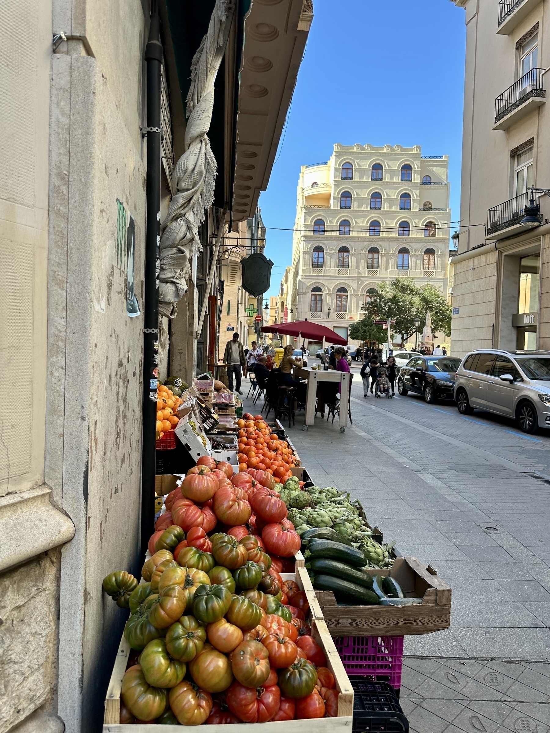 Outdoor vegetable stand on a narrow street. A restaurant patio full of guests is in the background.
