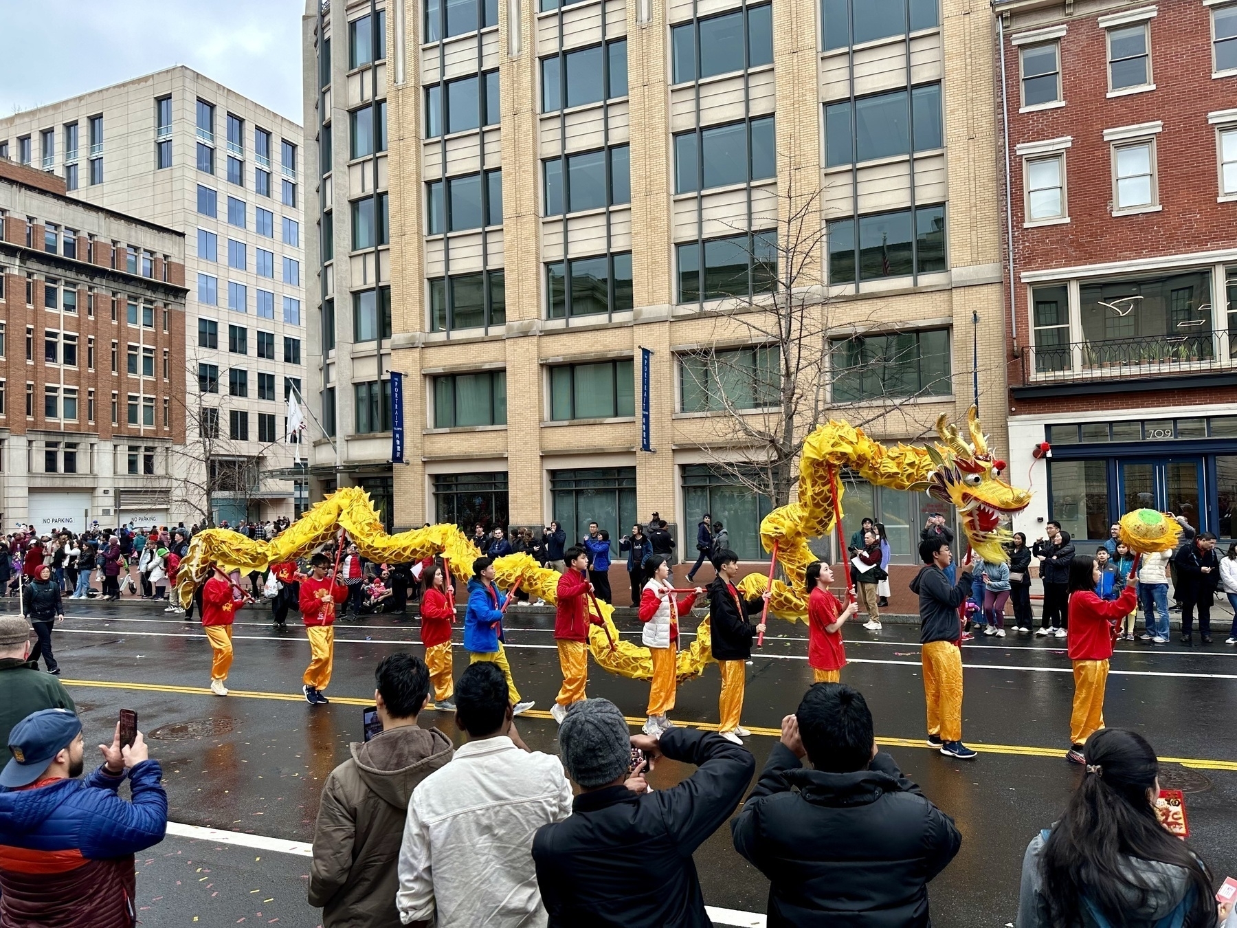 A long yellow Chinese dragon puppet carried by 9 people down a wet street, with spectators looking from both sides.