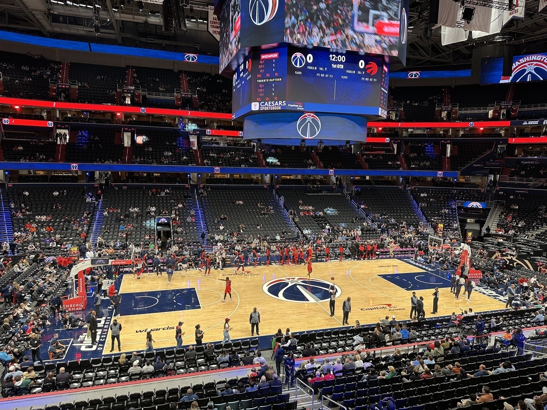 A basketball court inside an arena is shown with players warming up and a scoreboard displaying team logos (Wizards and Raptors)