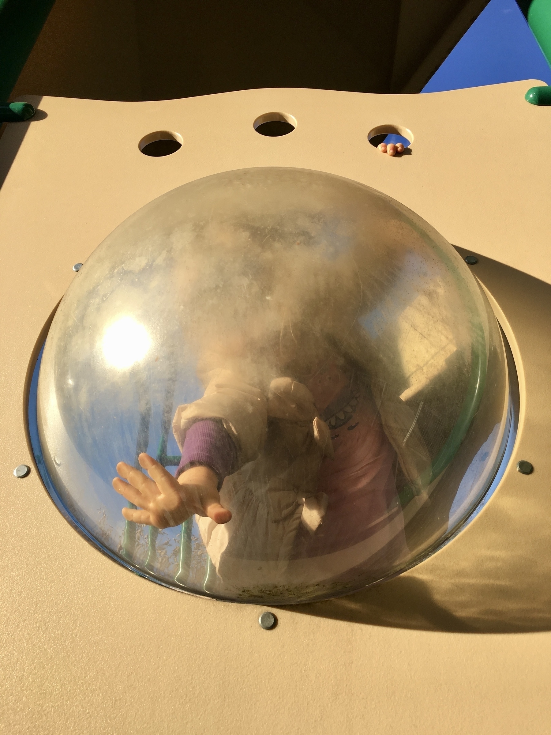 A toddler looking through a large opaque plastic bubble that’s part of a playground play set.