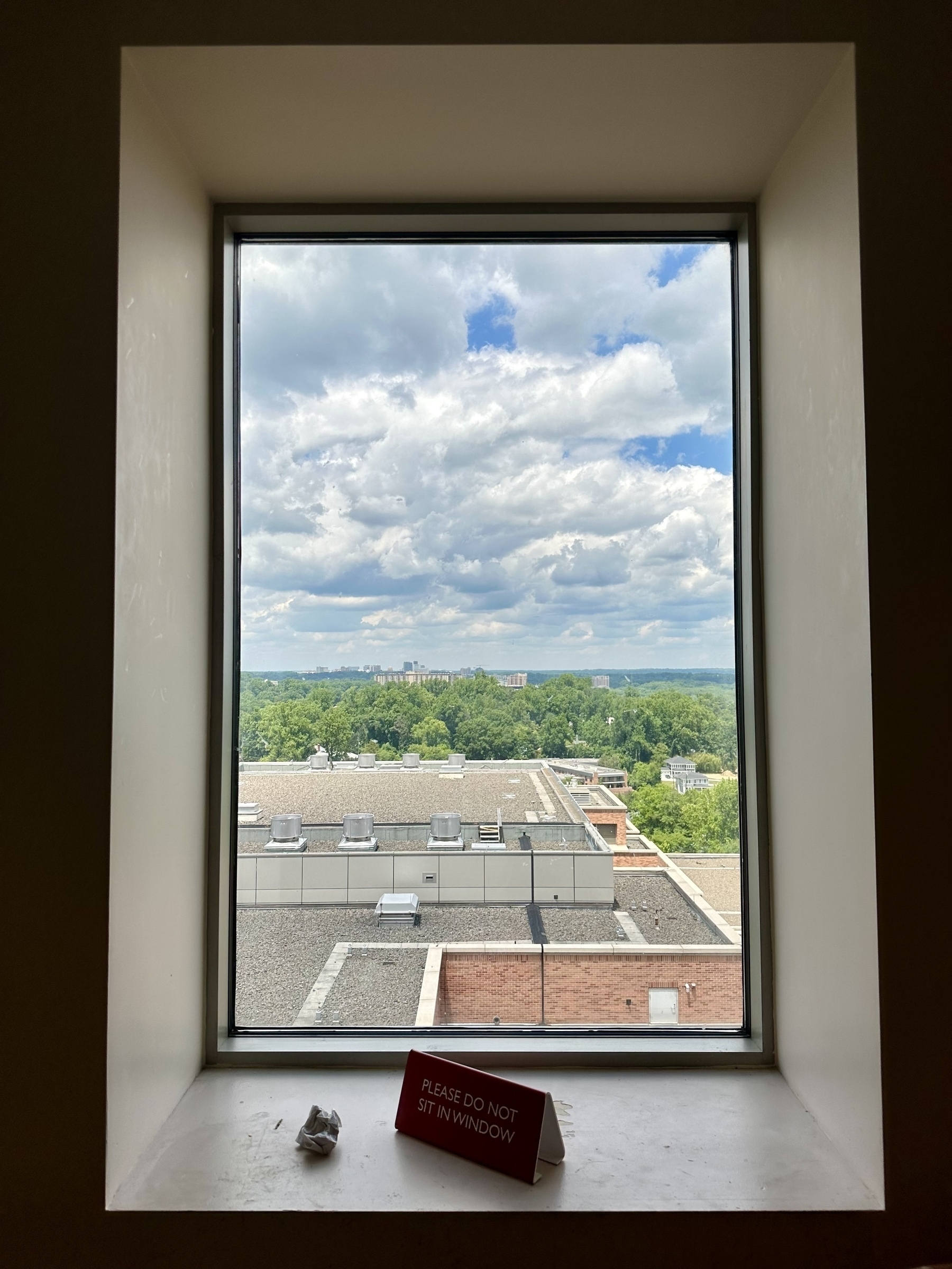 A window offers a view of an outside landscape with trees, buildings, and a partly cloudy sky, while a sign on the windowsill warns "PLEASE DO NOT SIT IN WINDOW."