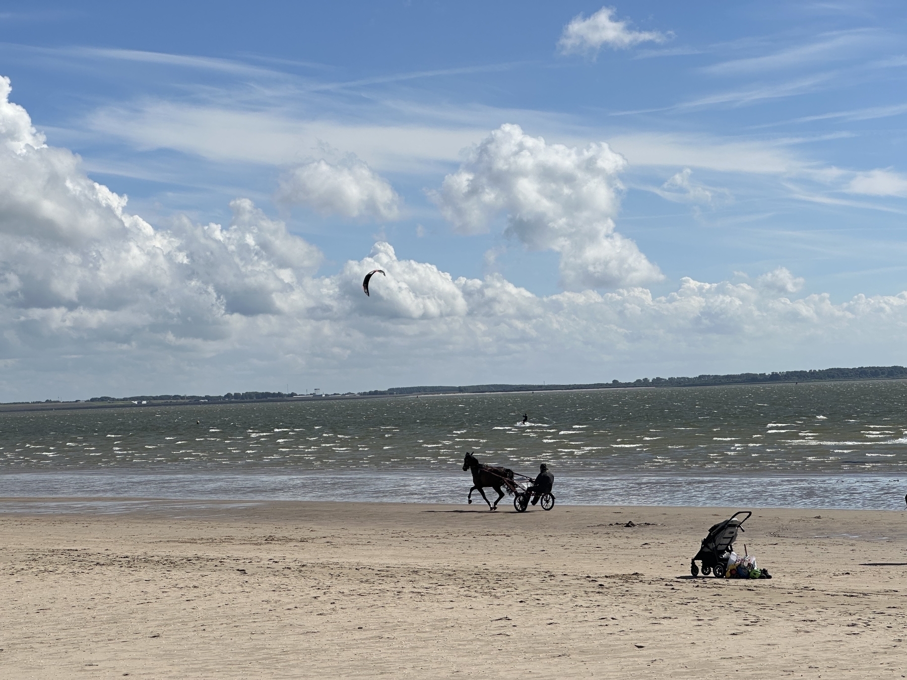 A horse pulling a carriage along a beach, with a baby stroller and some items in the sand, while a person kite surfing can be seen in the background.