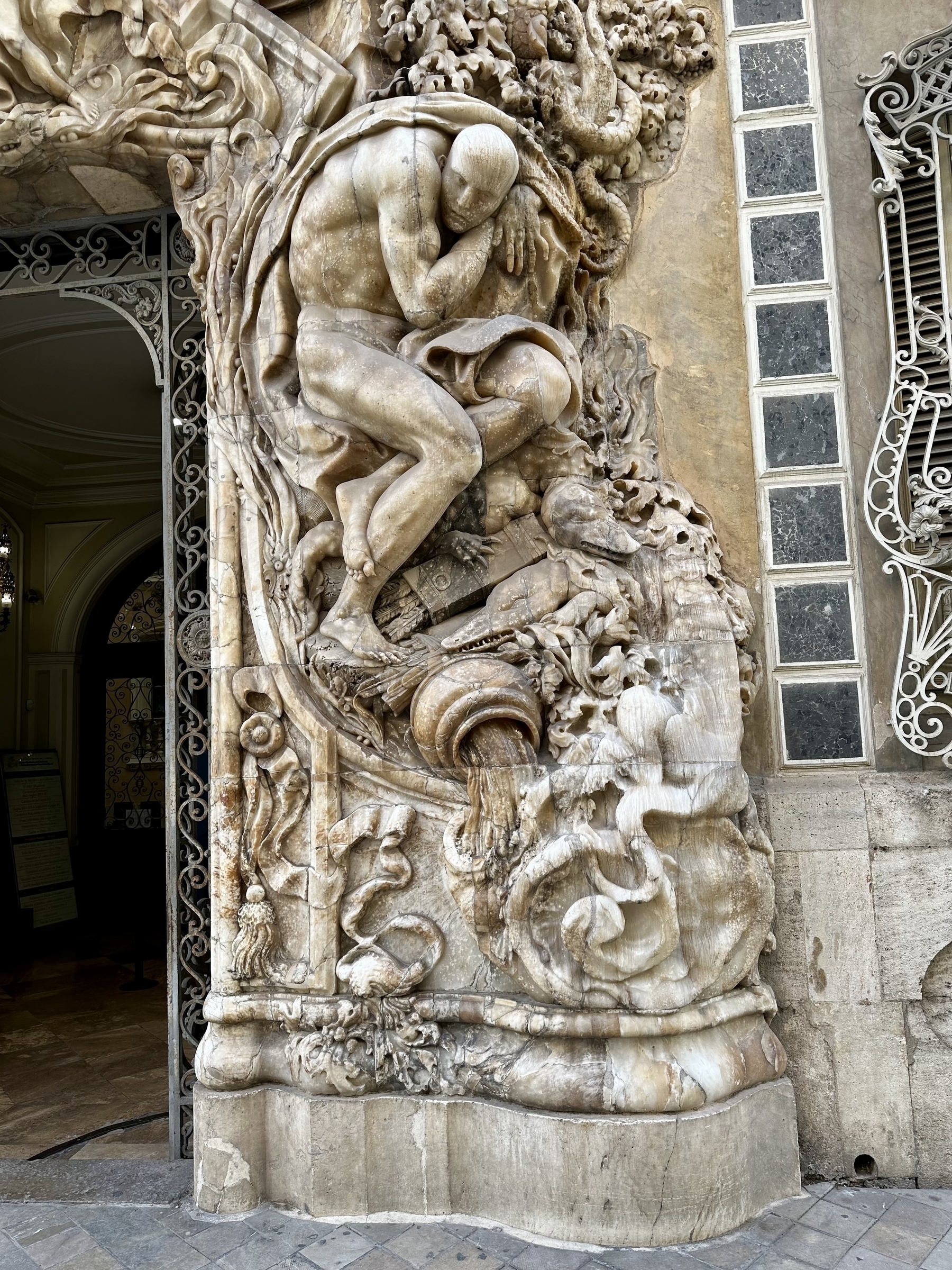 Building facade with a marble relief of a hunched man standing above a jug of water and an alligator.