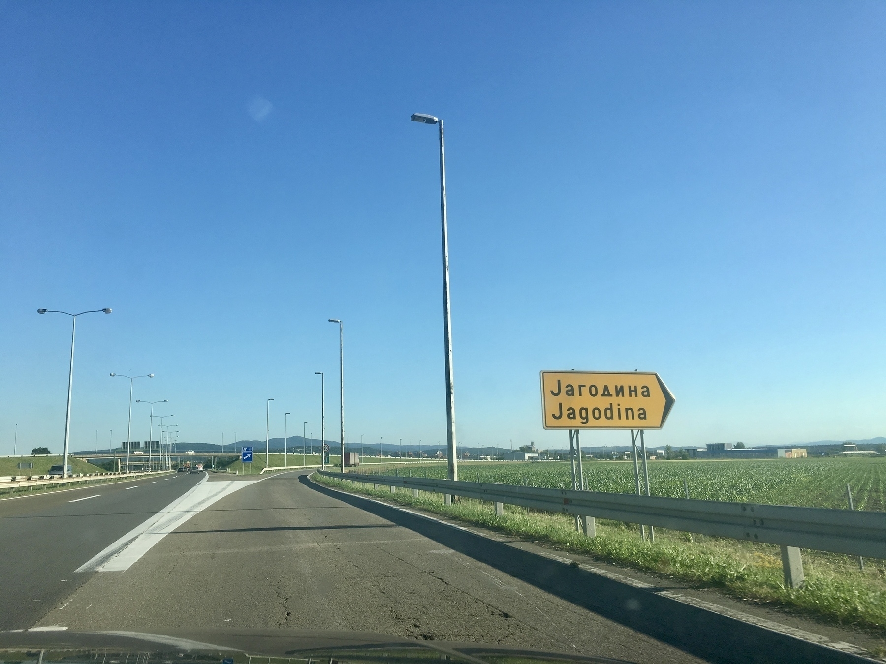 View from the windshield of a car taking an exit off a highway. A yellow sign with “Jagodina” written in Cyrillic and Latin alphabets points to the right towards a farm field.