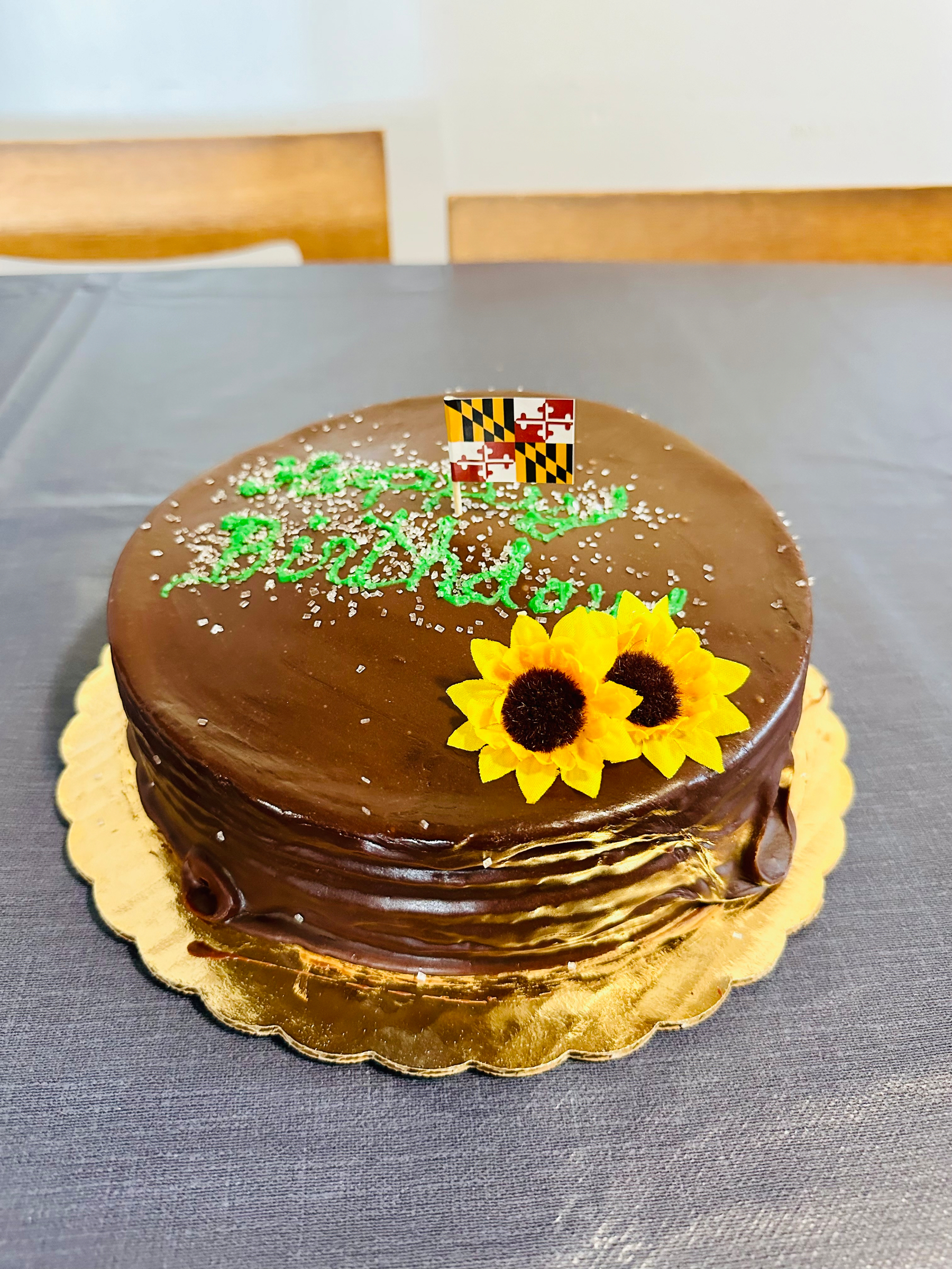 A chocolate cake decorated with yellow sunflower icing and a small flag on top sits on a gold base.