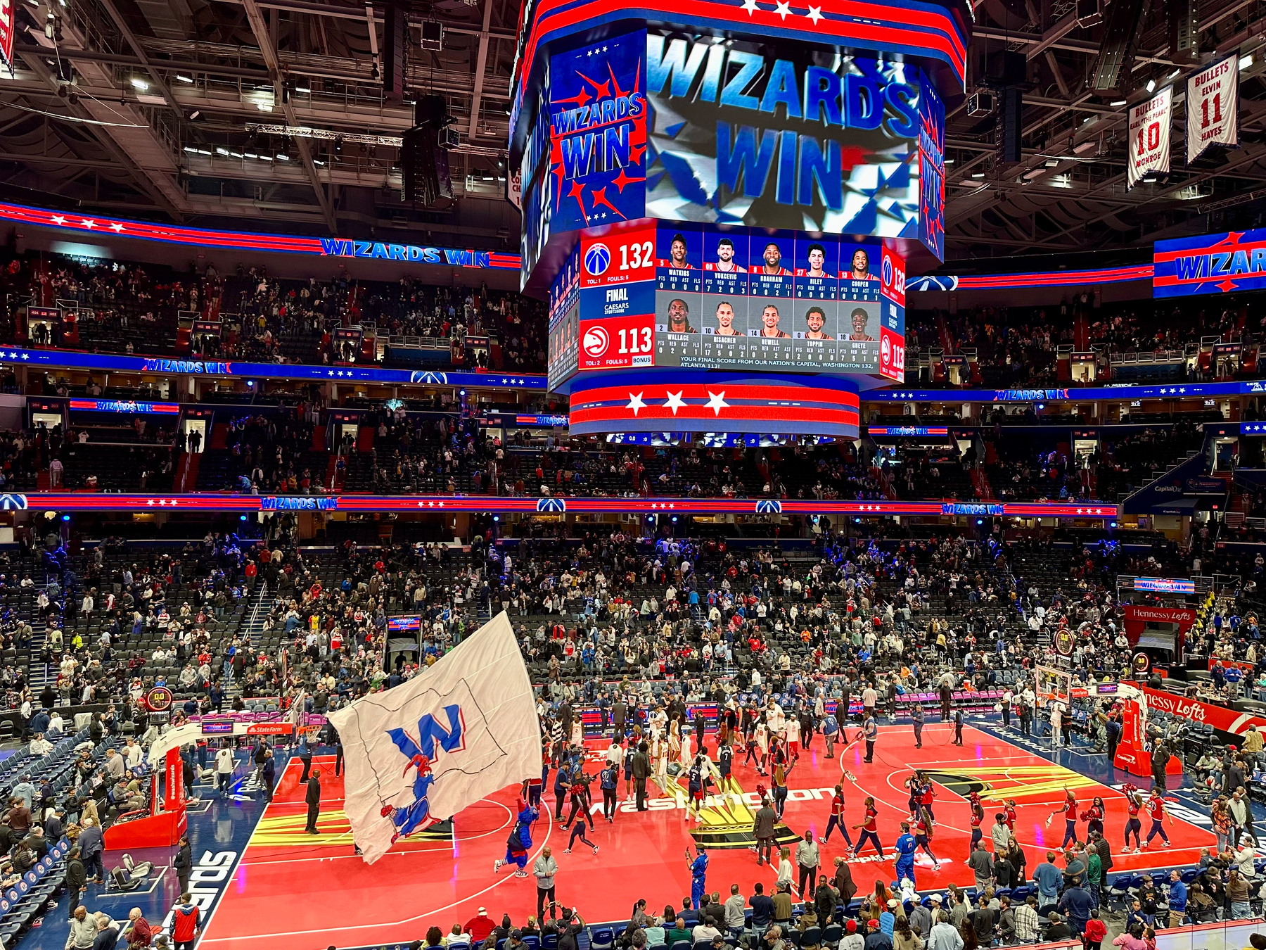 A basketball court celebrates a 132-113 Washington Wizards victory against the Atlanta Hawks, with fans and players on the floor and a large WIZARDS WIN display on the scoreboard.