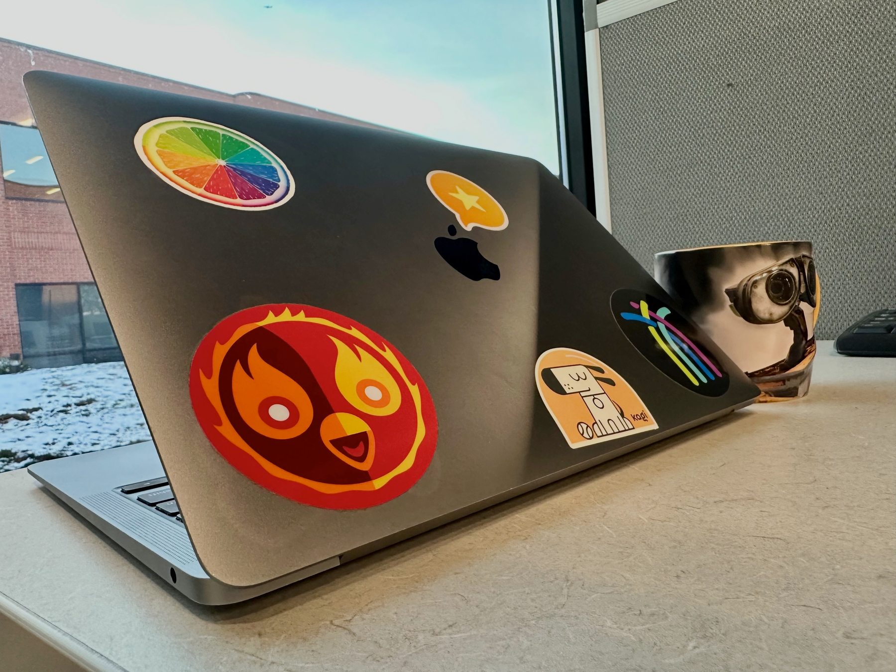 A laptop adorned with colorful stickers is positioned on a desk next to a mug featuring an image of WALL-E, with a snowy view outside the window.