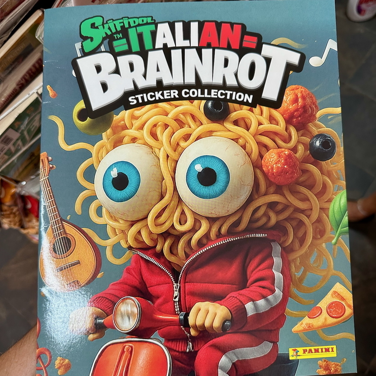 AI-generated image of a cartoon character with a spaghetti head, large blue eyes, and a red tracksuit rides a Vespa scooter, surrounded by playful elements like a guitar, meatballs, and musical notes. The title above is “Italian Brainrot Sticker Collection”.