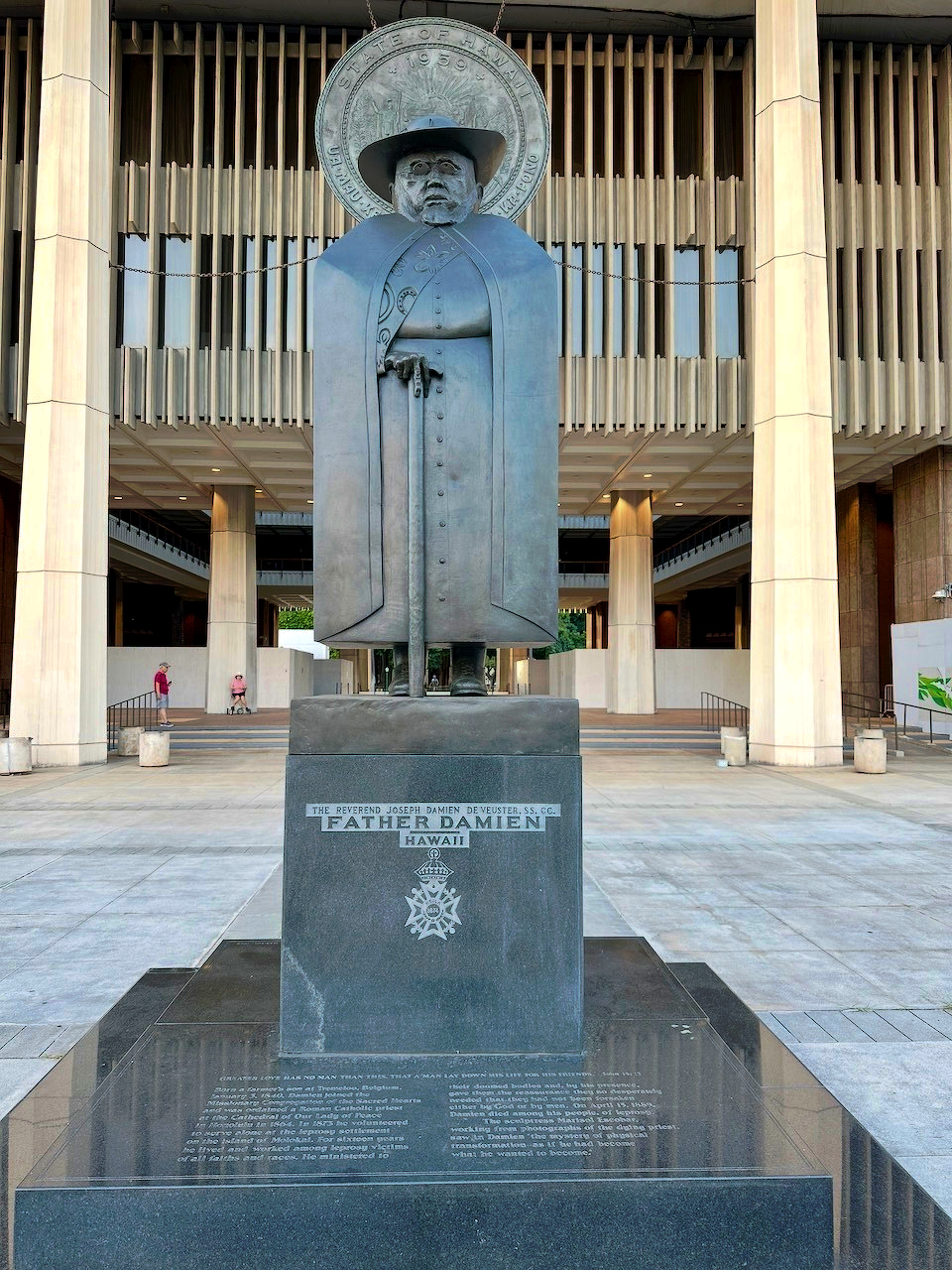 A bronze statue of Father Damien stands prominently on a pedestal in front of a modern building with tall columns.