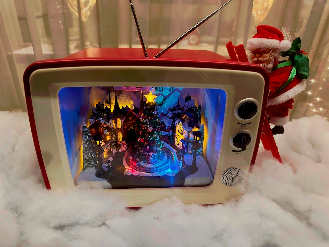 A vintage television with a Christmas village scene inside is surrounded by snow-like material, with a Santa figure climbing a ladder.