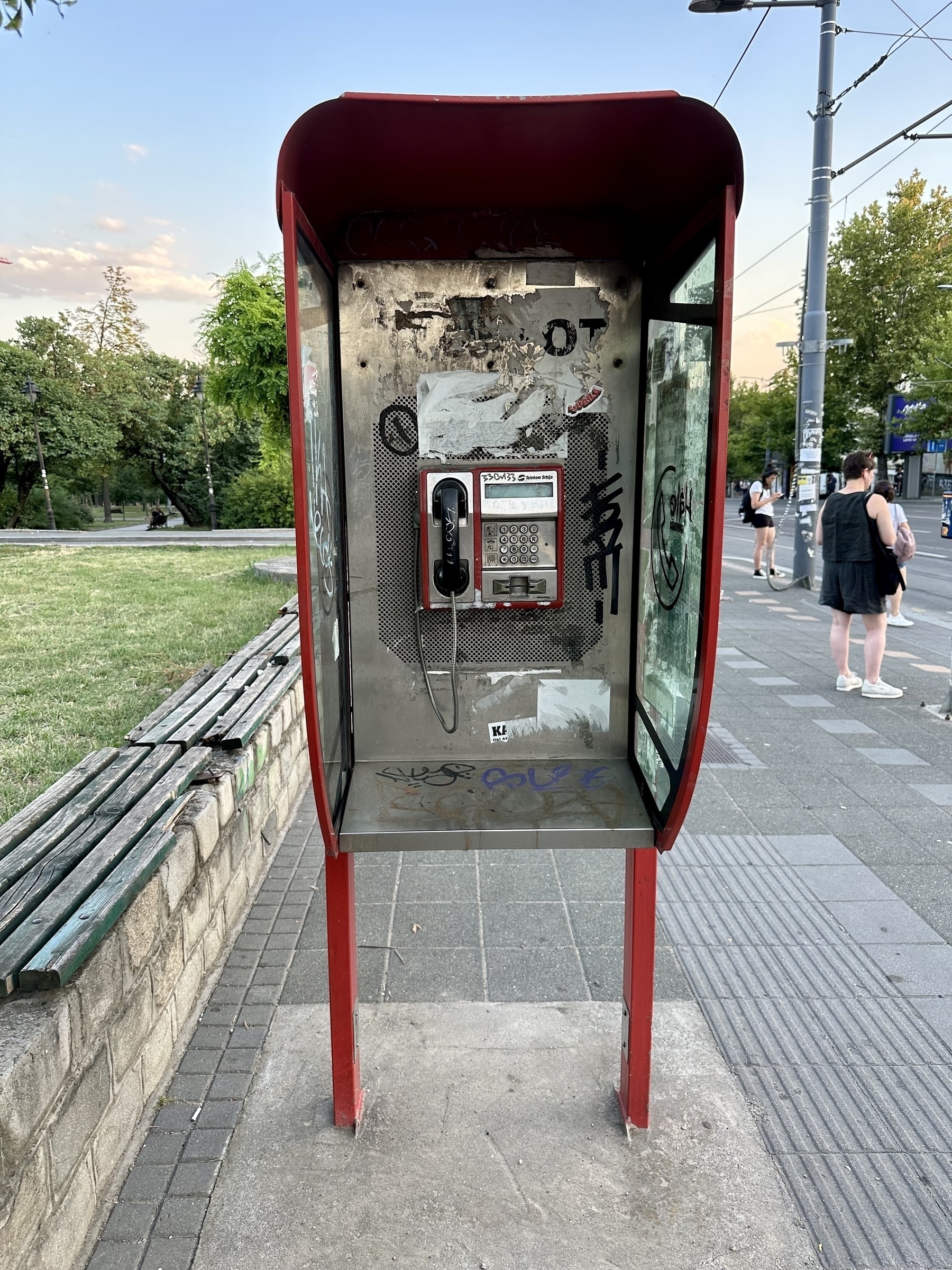 A red phone booth with a public payphone stands on a sidewalk, surrounded by green trees and pedestrians.