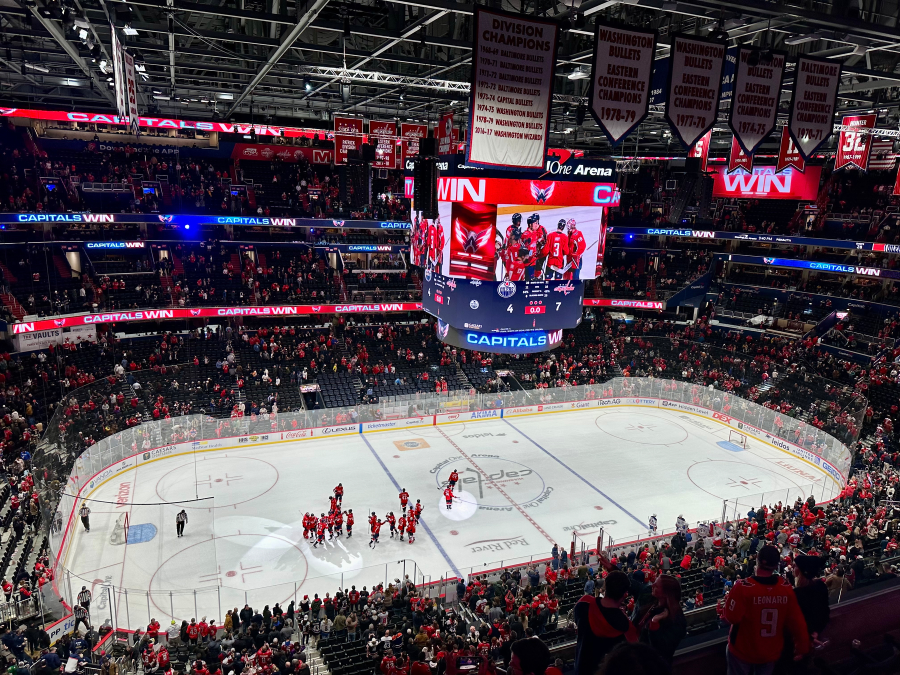 A hockey arena filled with fans celebrates a win with players on the ice and a scoreboard displaying the victory of the Washington Capitals against the Edmonton Oilers.