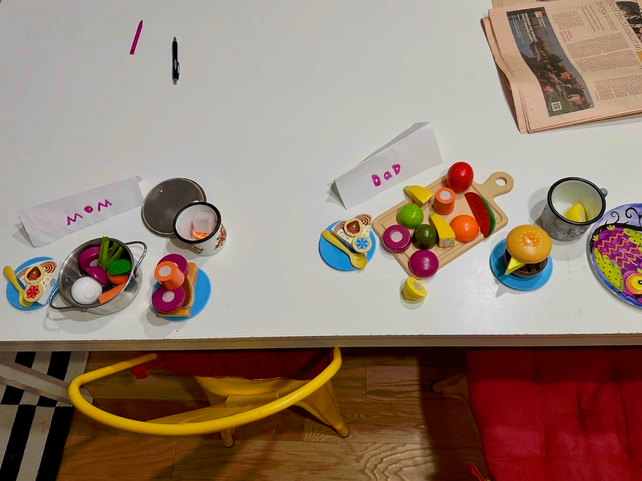 Two table settings feature toy food and name cards labeled Mom and Dad, with a newspaper and pen nearby.