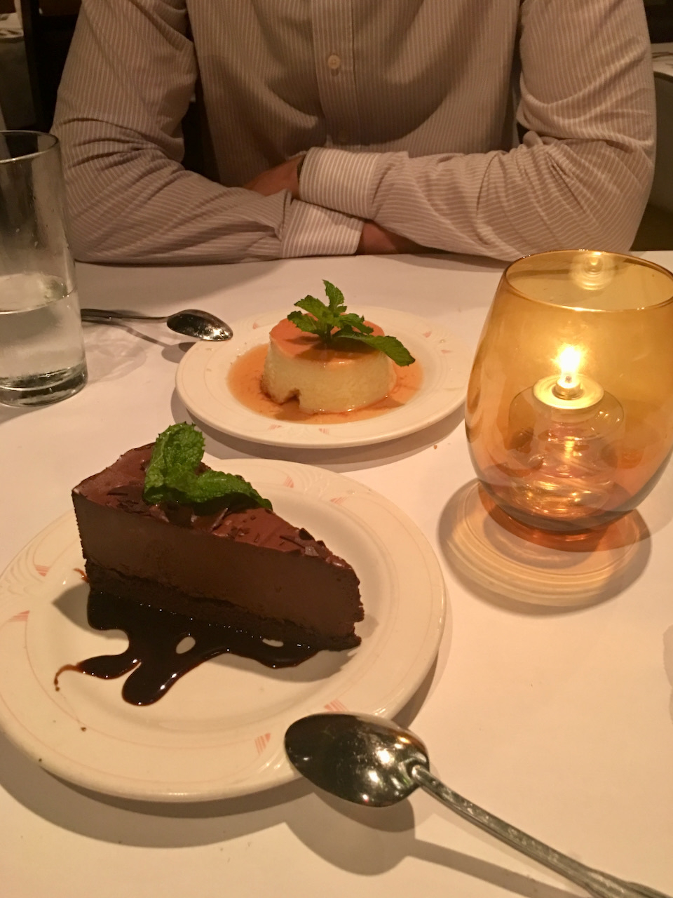 A person is seated at a table with chocolate cake and flan, accompanied by a candle and a glass of water.