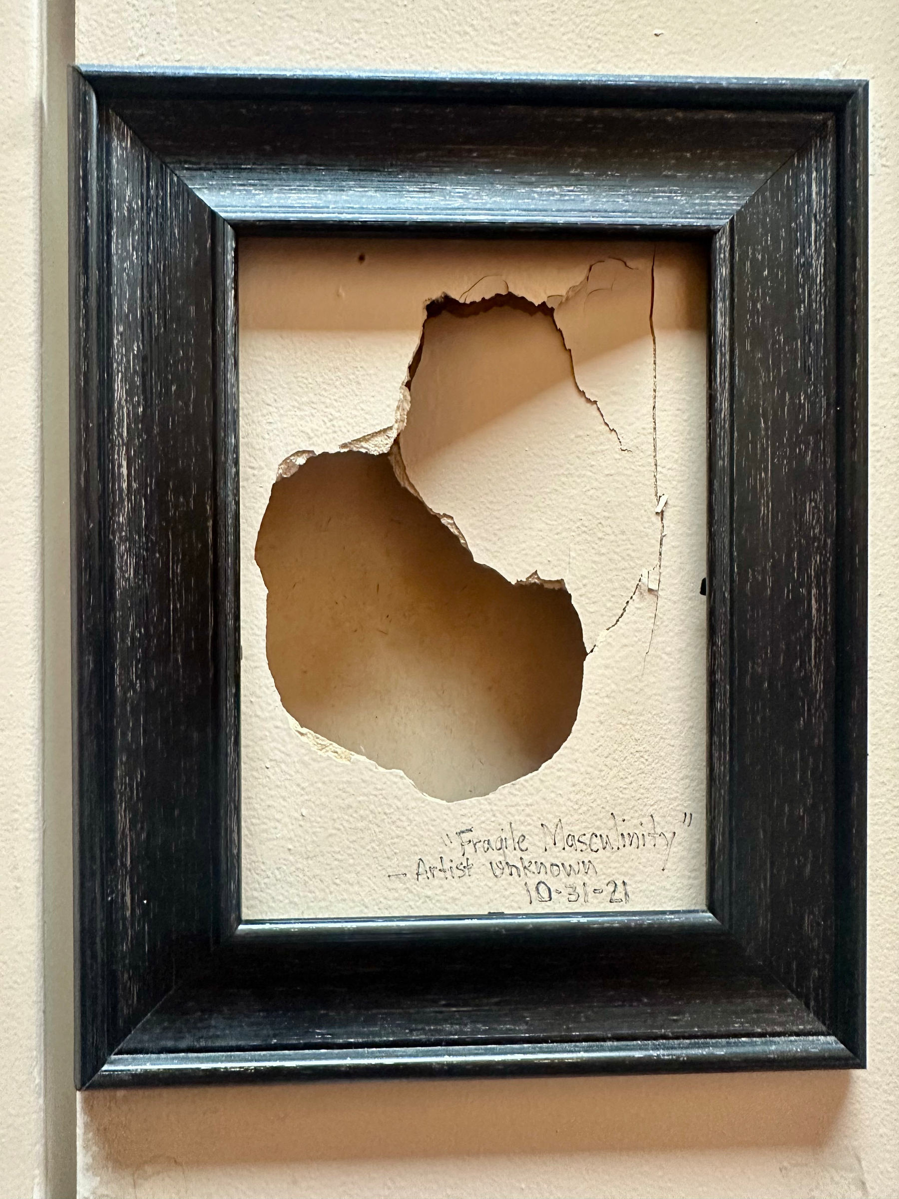 A black frame encases a broken section of drywall, titled Fragile Masculinity with a note from an anonymous artist dated 10-31-21.