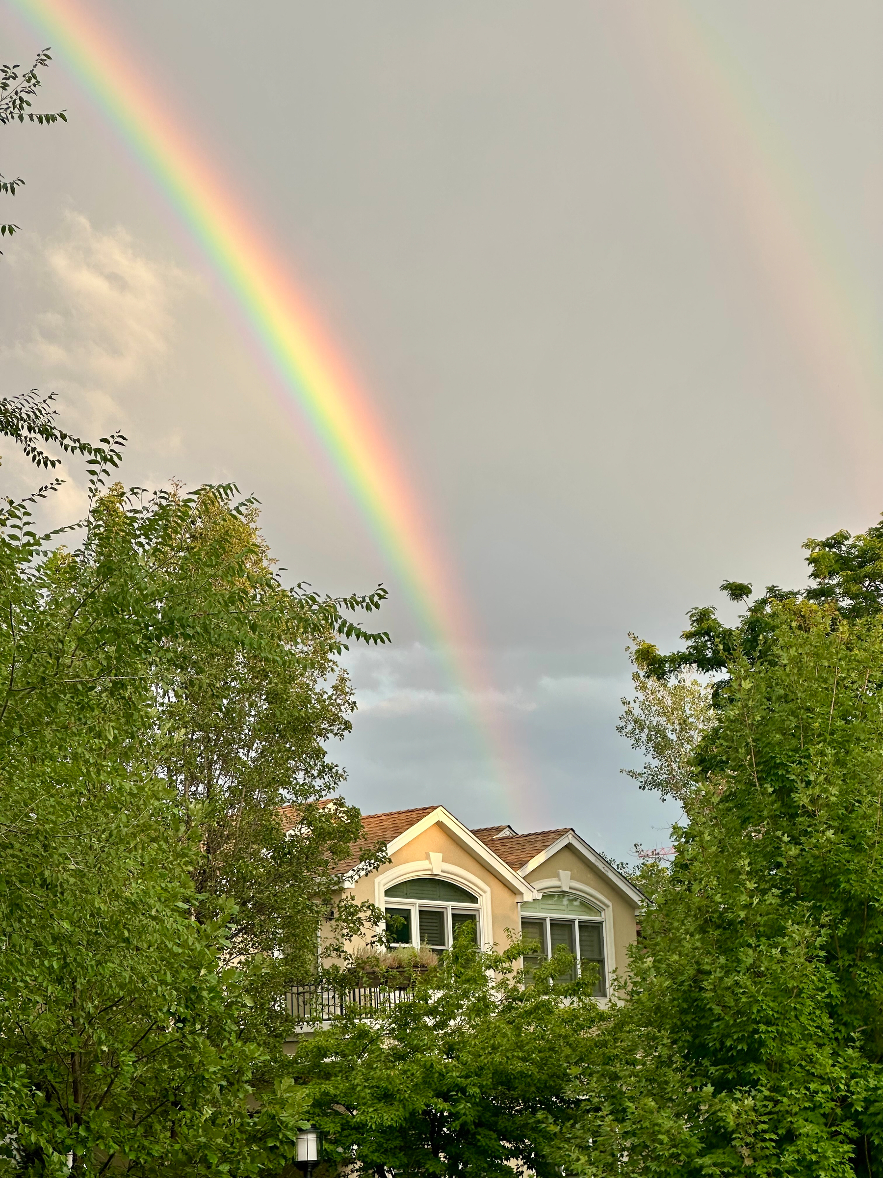 A vibrant rainbow arches across a cloudy sky above a house partially obscured by lush green trees.