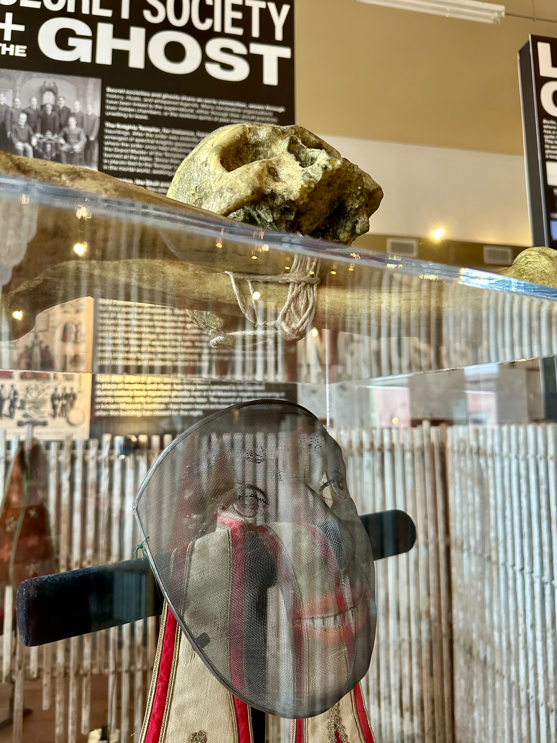 A vintage fencing mask used in Odd Fellow rituals is displayed in a museum setting along with historical information about the connection between ghosts and secret societies.