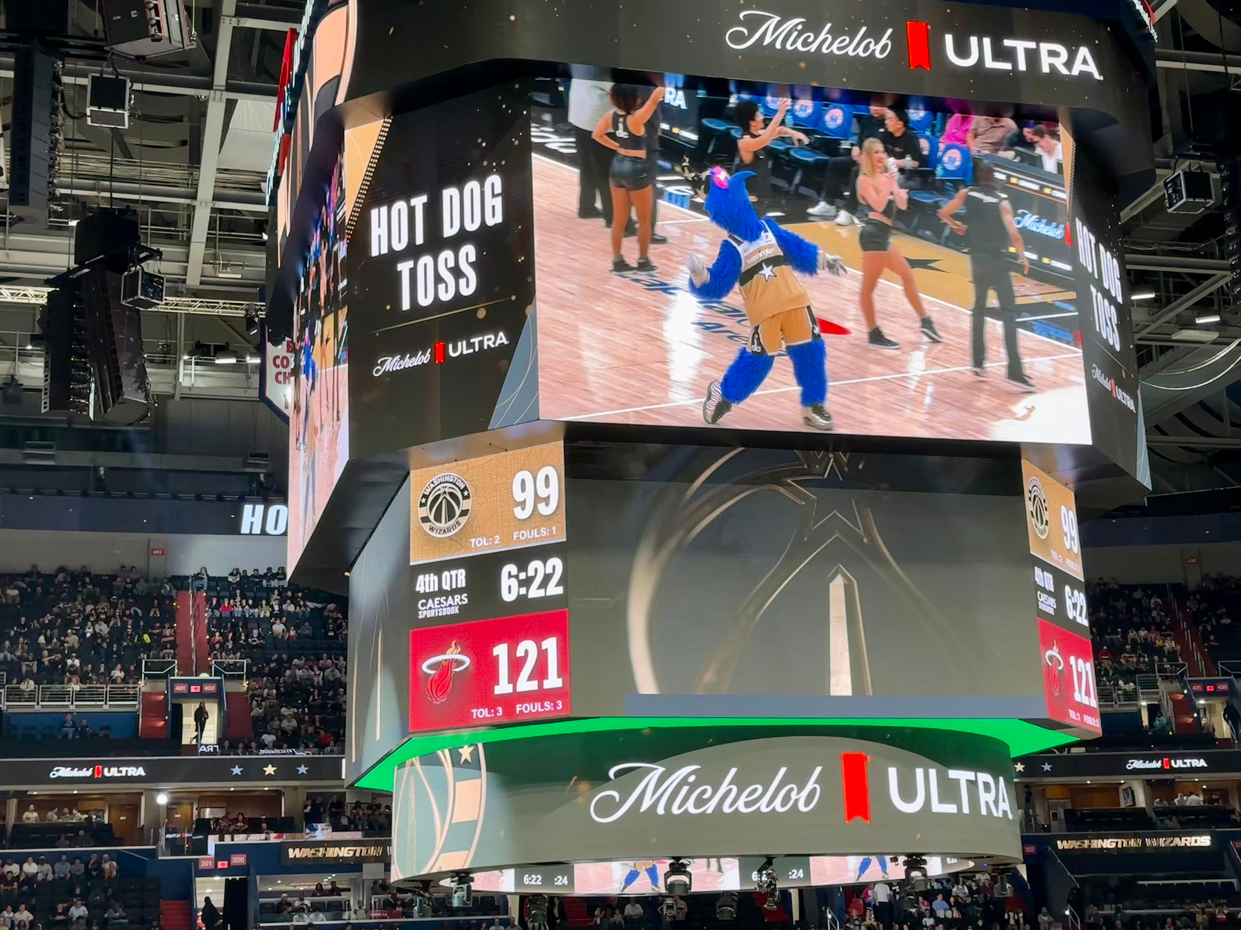 A scoreboard displays a basketball game score of 121-99, with a dance or halftime activity featuring a mascot throwing a hot dog shown on the large screens above the court.