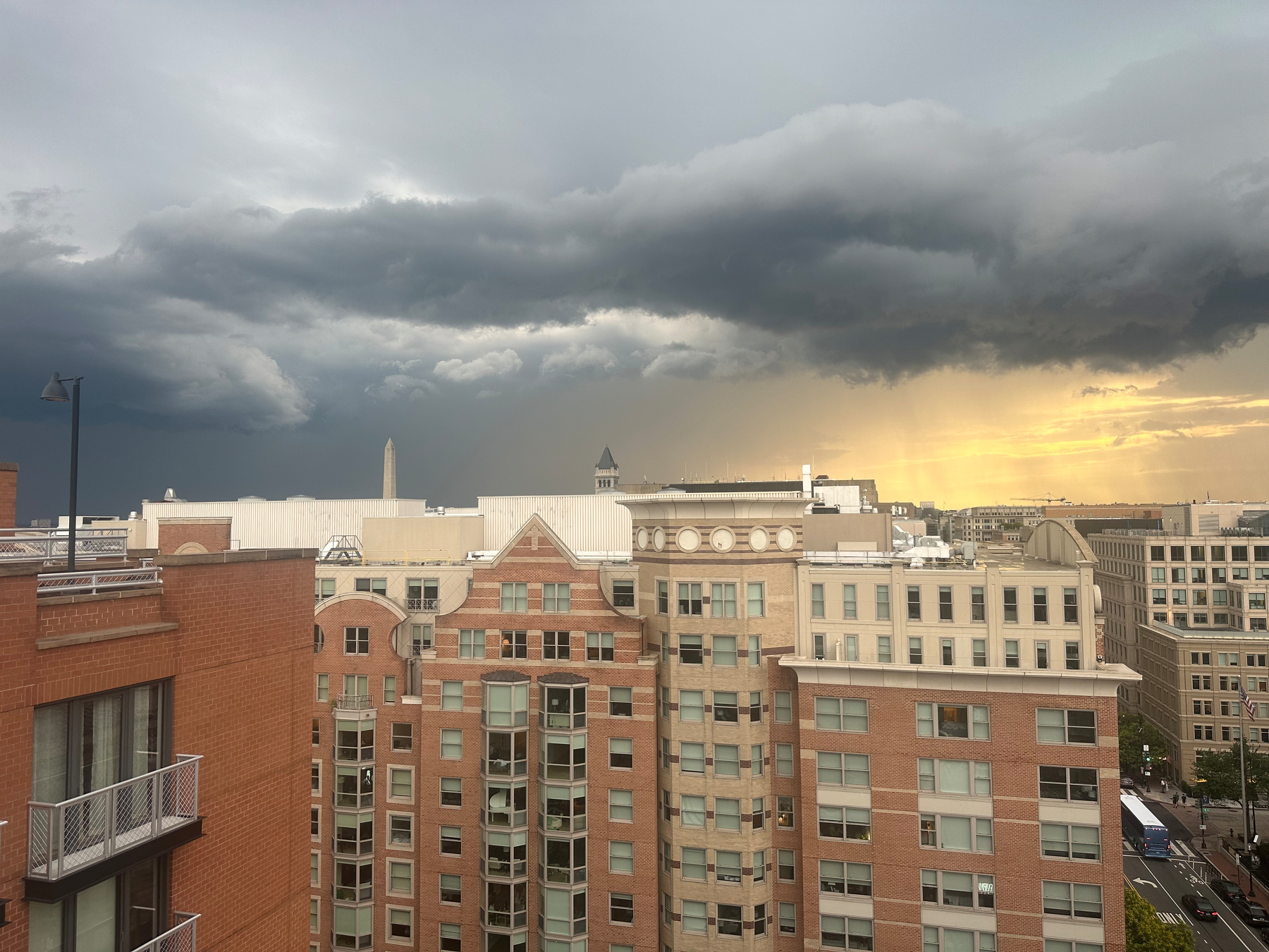A cityscape with tall buildings and the Washington monument is set against a dramatic sky with dark storm clouds and a golden sunset in the distance.