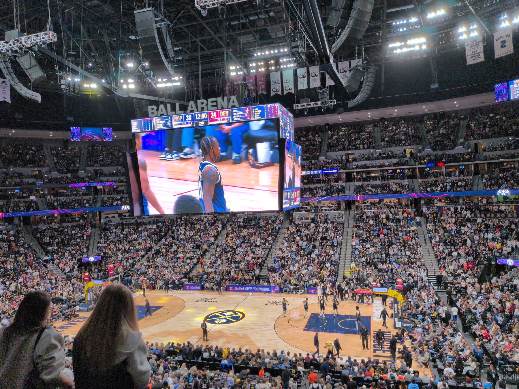 A basketball game is taking place in a large indoor arena filled with spectators, with a player shown on the central scoreboard. Nuggets as losing to the Pelicans 38-24 at the beginning of the second quarter.