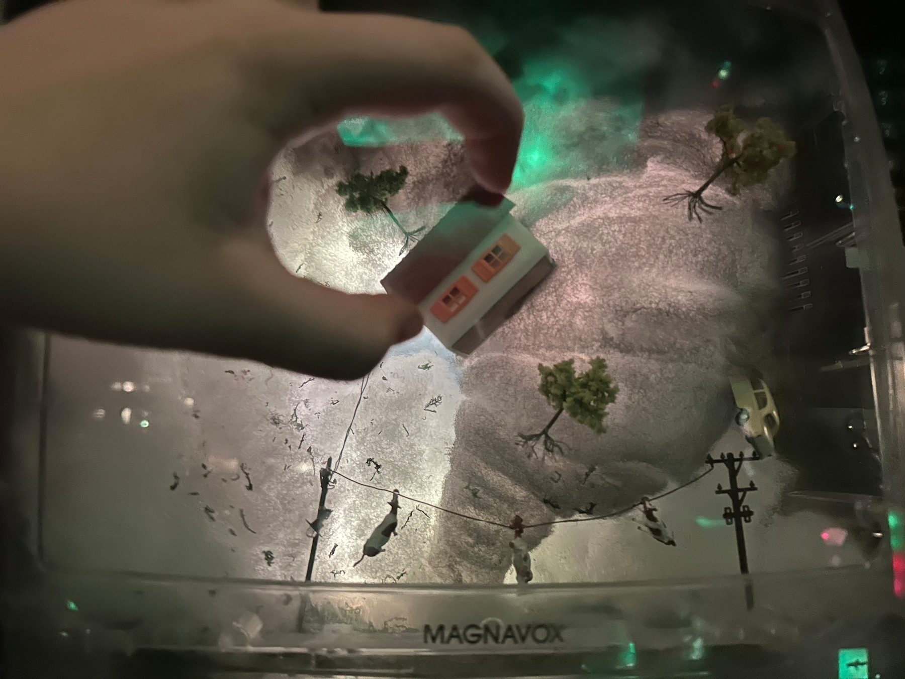 A hand is holding a small house above a miniature scene labeled MAGNAVOX that resembles a tornado touching the ground with trees and utility poles around it.