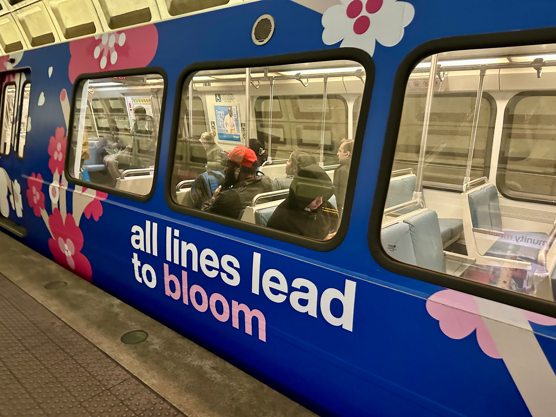 A brightly decorated metro train with floral patterns and the phrase “All lines lead to bloom” is seen with passengers inside.