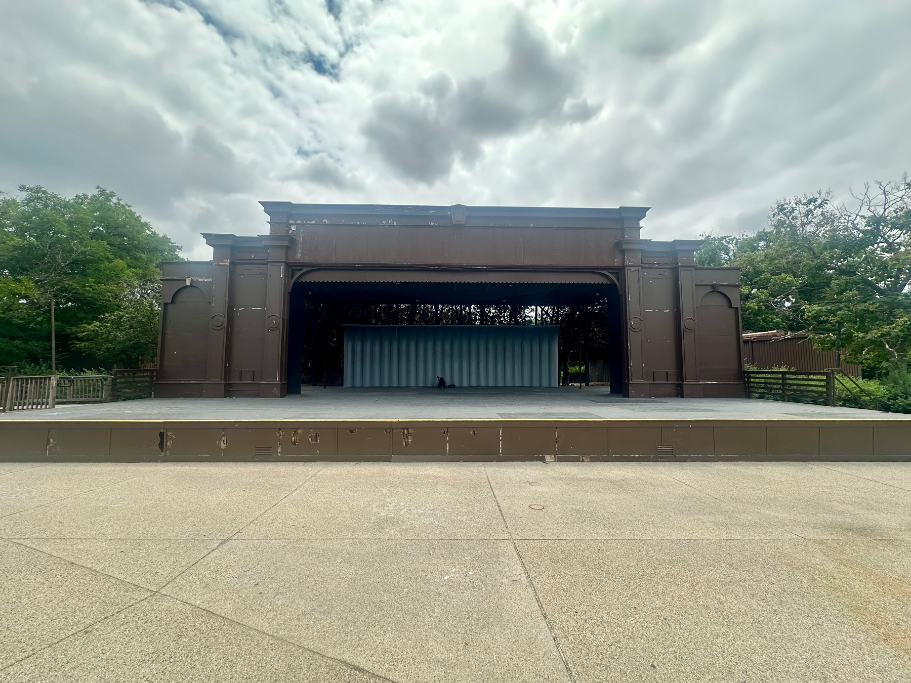 An empty, tattered outdoor stage with a covered backdrop stands under a cloudy sky surrounded by trees.