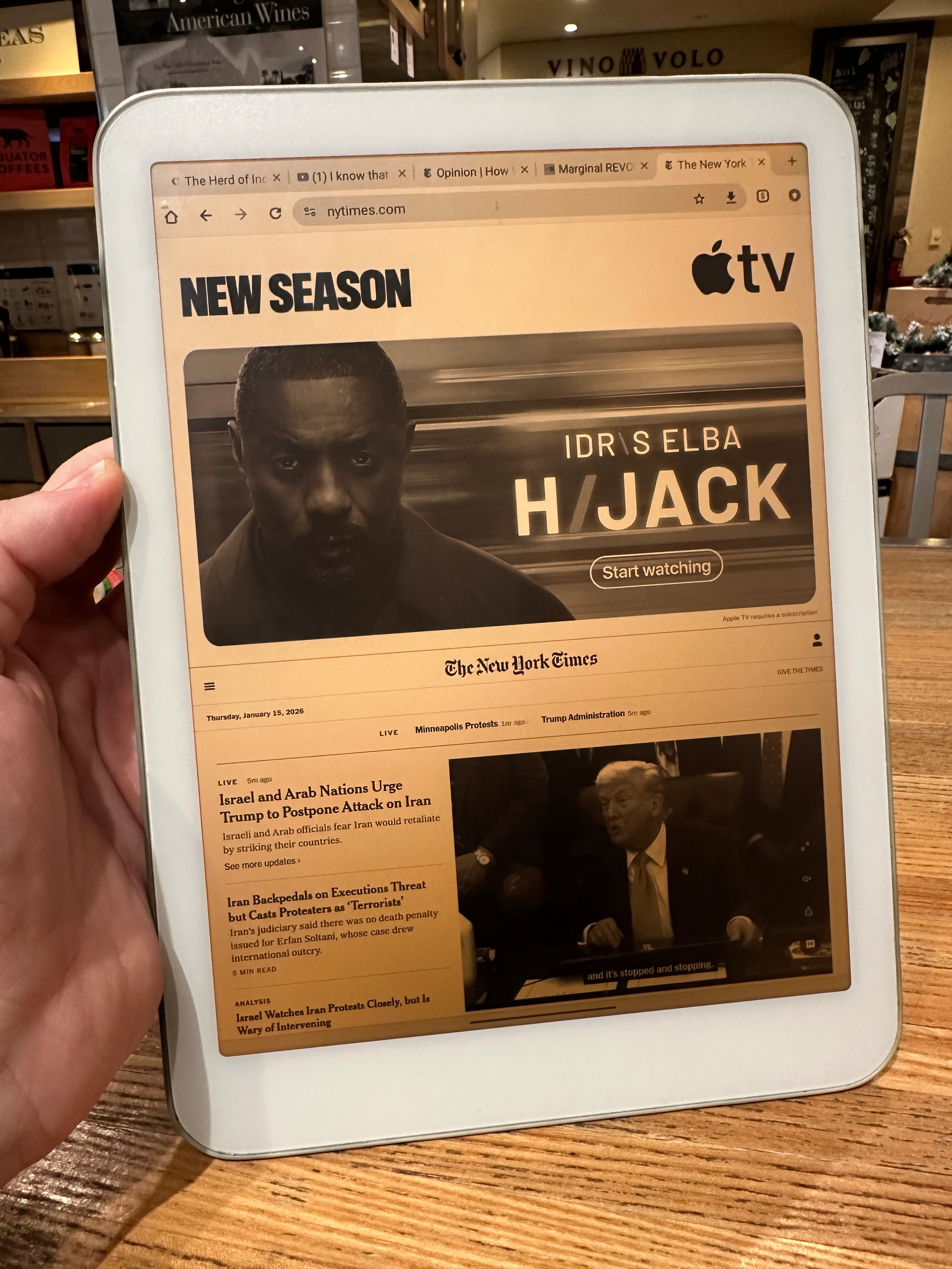 A person holds a tablet displaying a digital newspaper (NYT) with a prominent Apple TV advertisement featuring Idris Elba for a show called Hijack. Less than an eight of the screen contains any news.