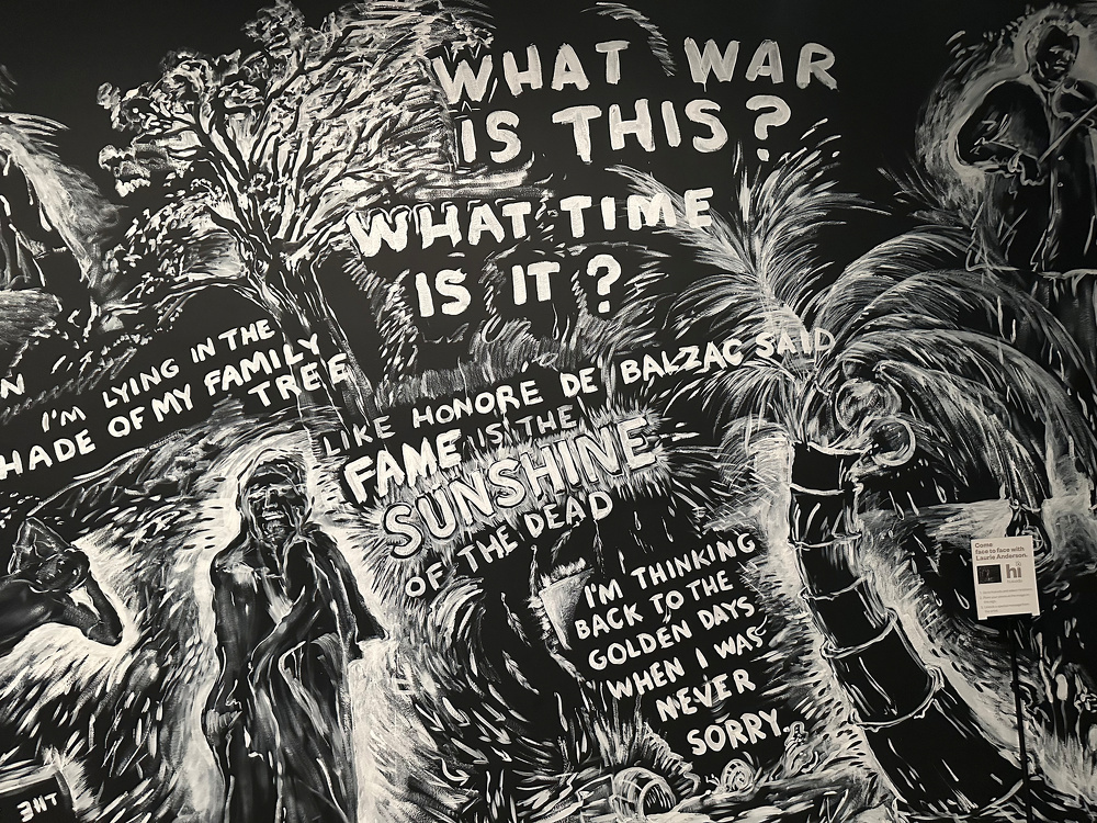 A black and white mural features abstract artwork with phrases like WHAT WAR IS THIS? and LIKE HONORE DE BALZAC SAID FAME IS THE SUNSHINE OF THE DEAD, surrounded by various figures and elements.