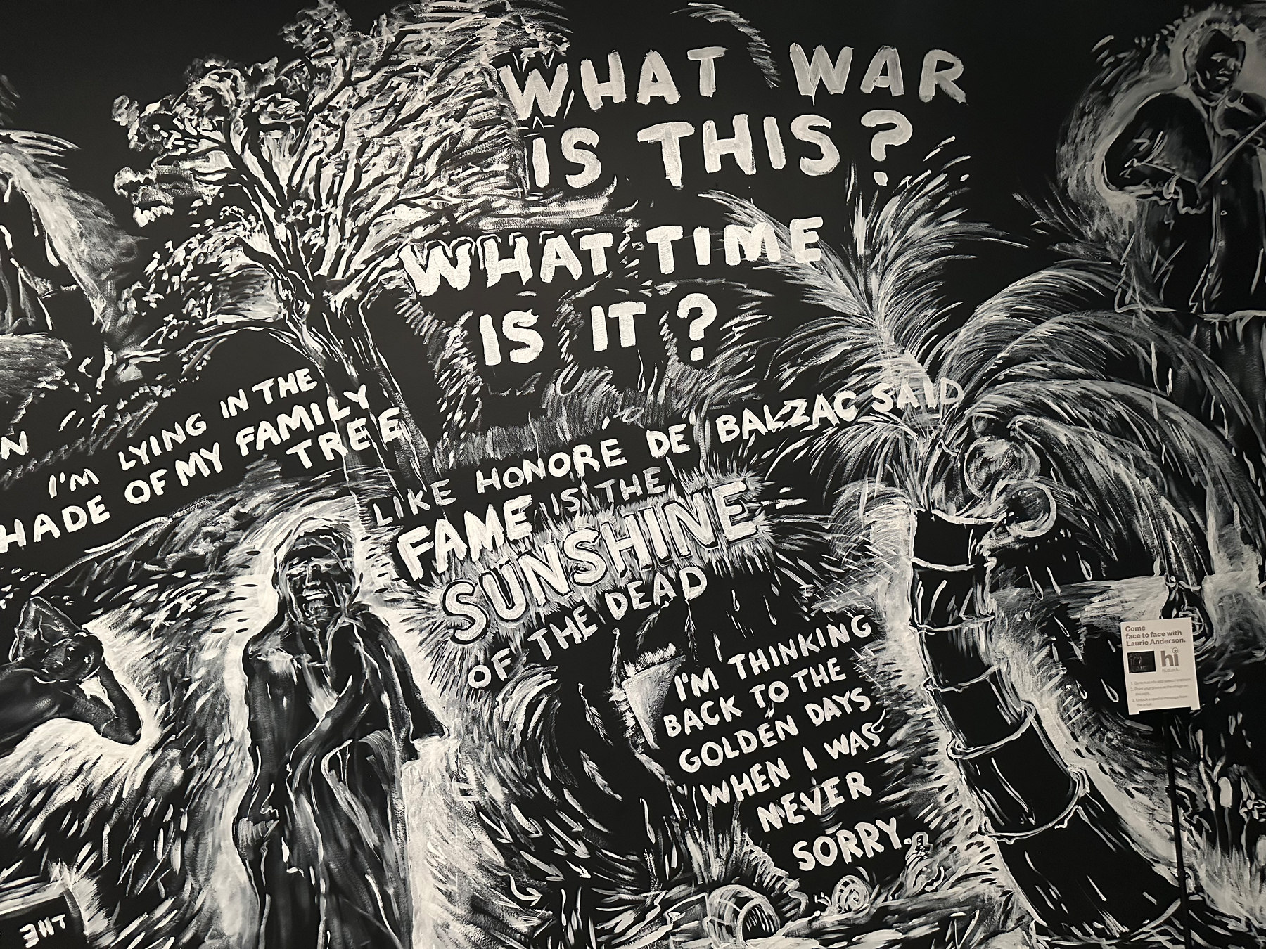 A black and white mural features abstract artwork with phrases like WHAT WAR IS THIS? and LIKE HONORE DE BALZAC SAID FAME IS THE SUNSHINE OF THE DEAD, surrounded by various figures and elements.