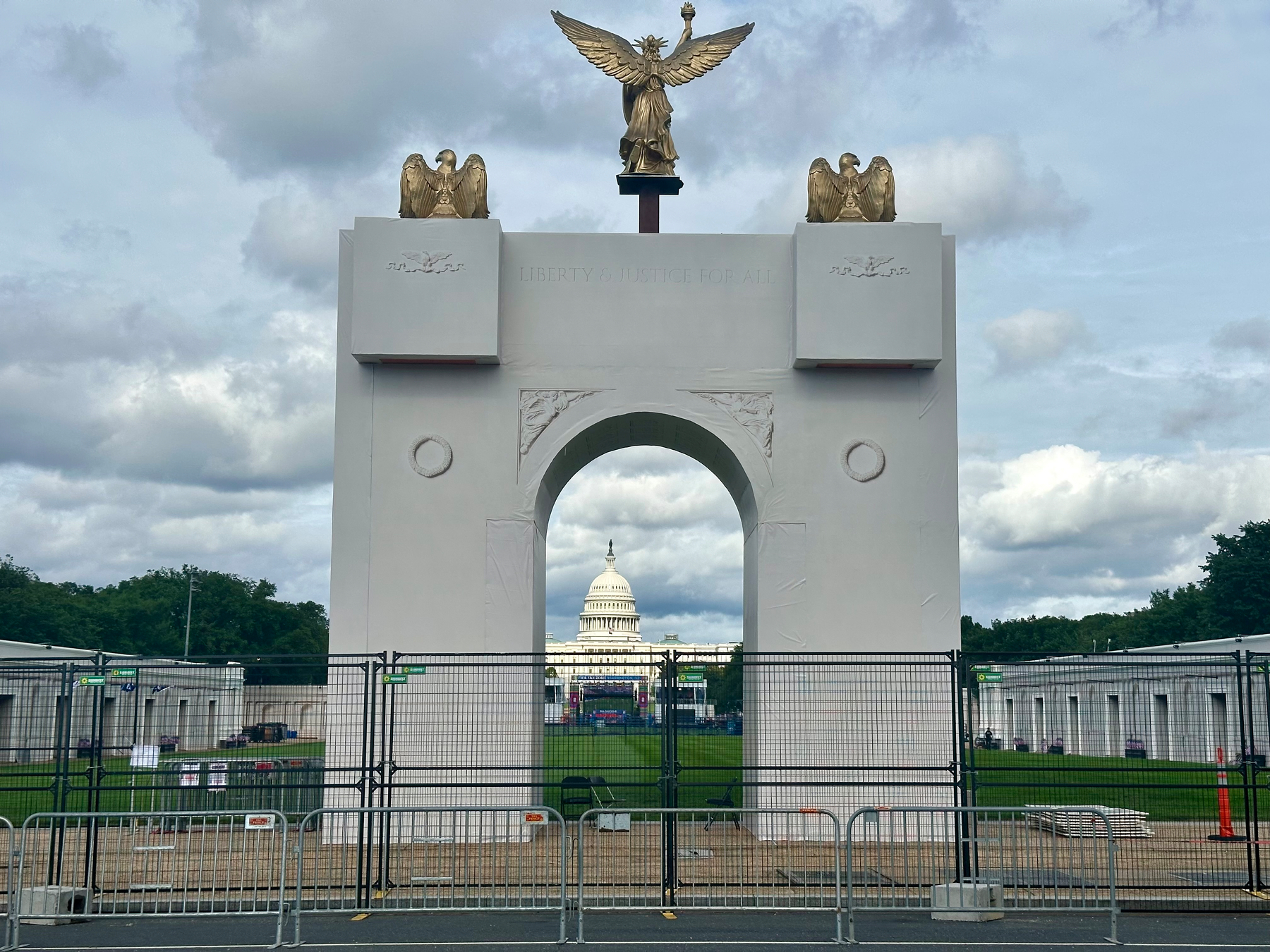 A large white memorial arch with golden eagle statues on top stands in front of the U.S. Capitol building, with fencing and barricades surrounding the area.