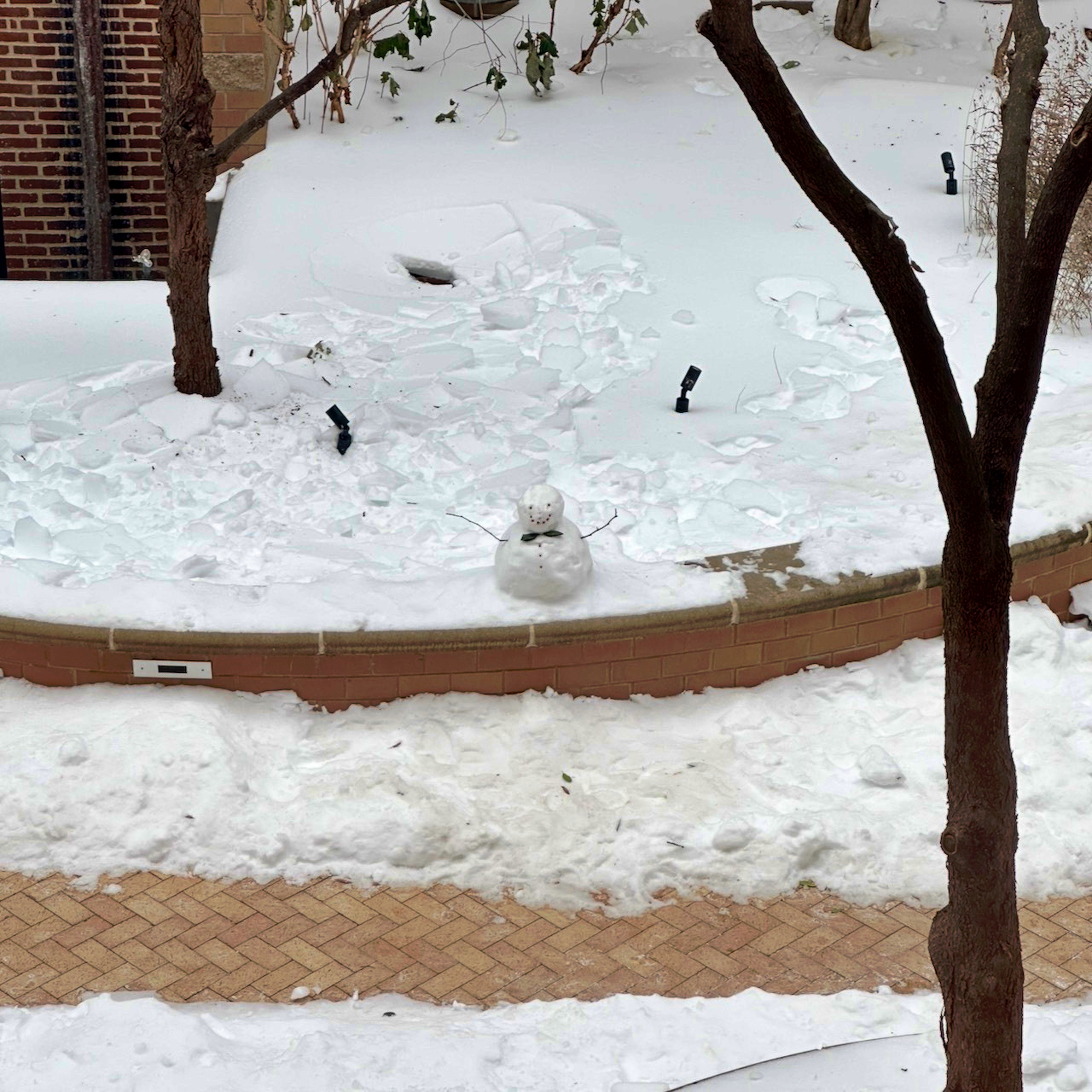 A small snowman with stick arms stands in a snowy area near a brick border and bare trees.