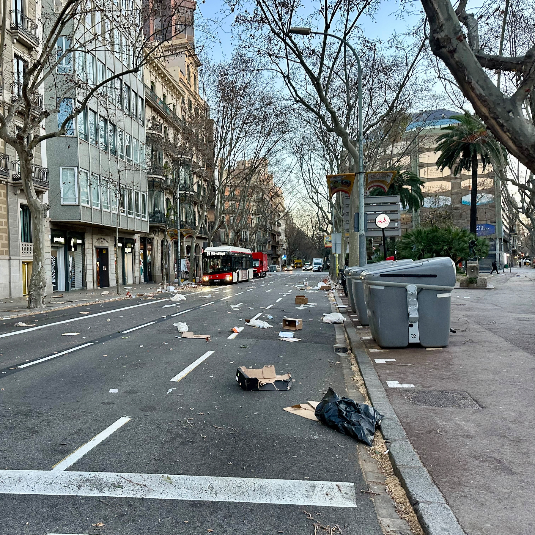 A city street shows litter scattered across the road with a bus parked along the sidewalk on a clear day.