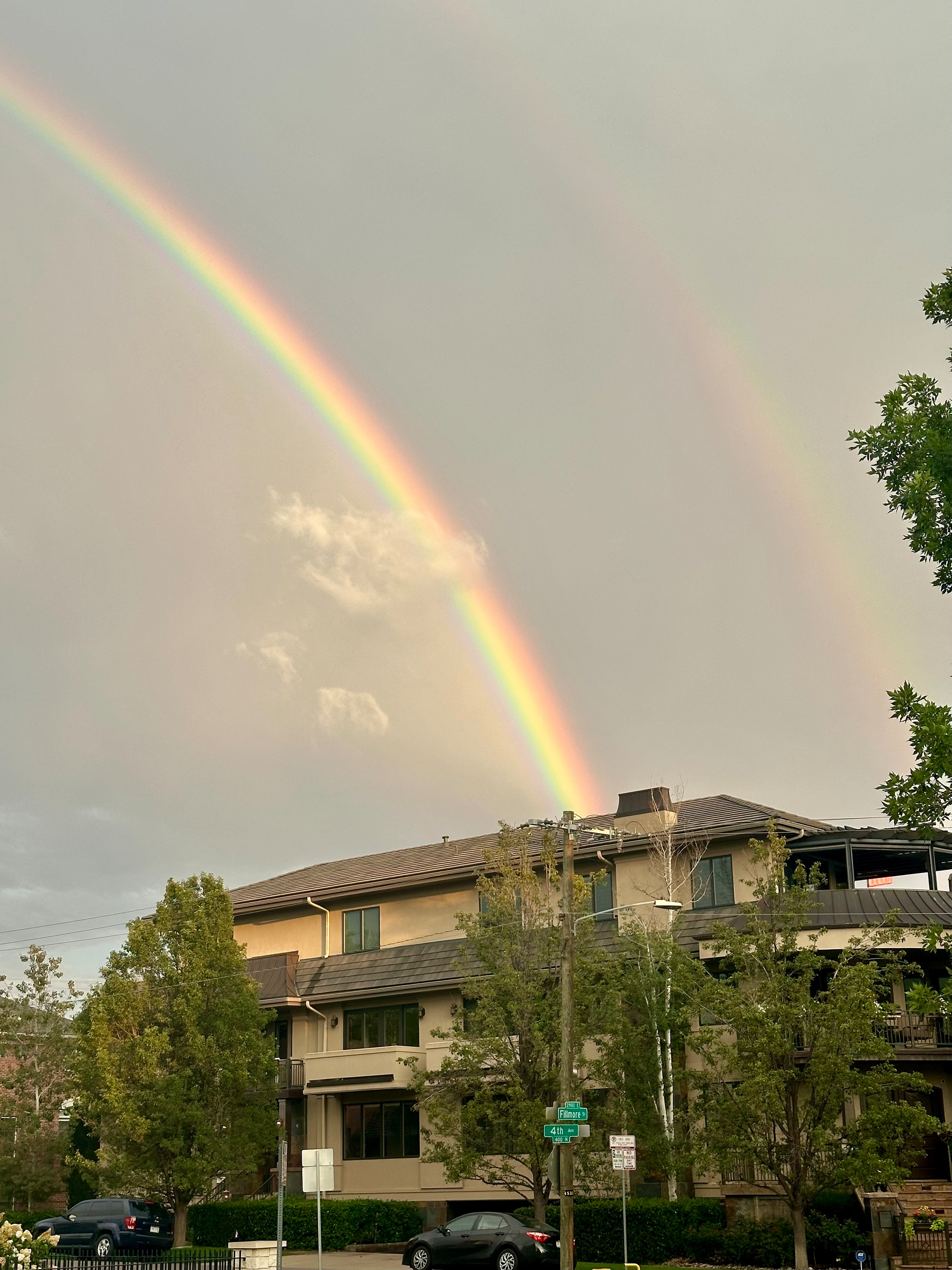 A vivid double rainbow arcs across the sky above a residential building surrounded by trees and parked cars.