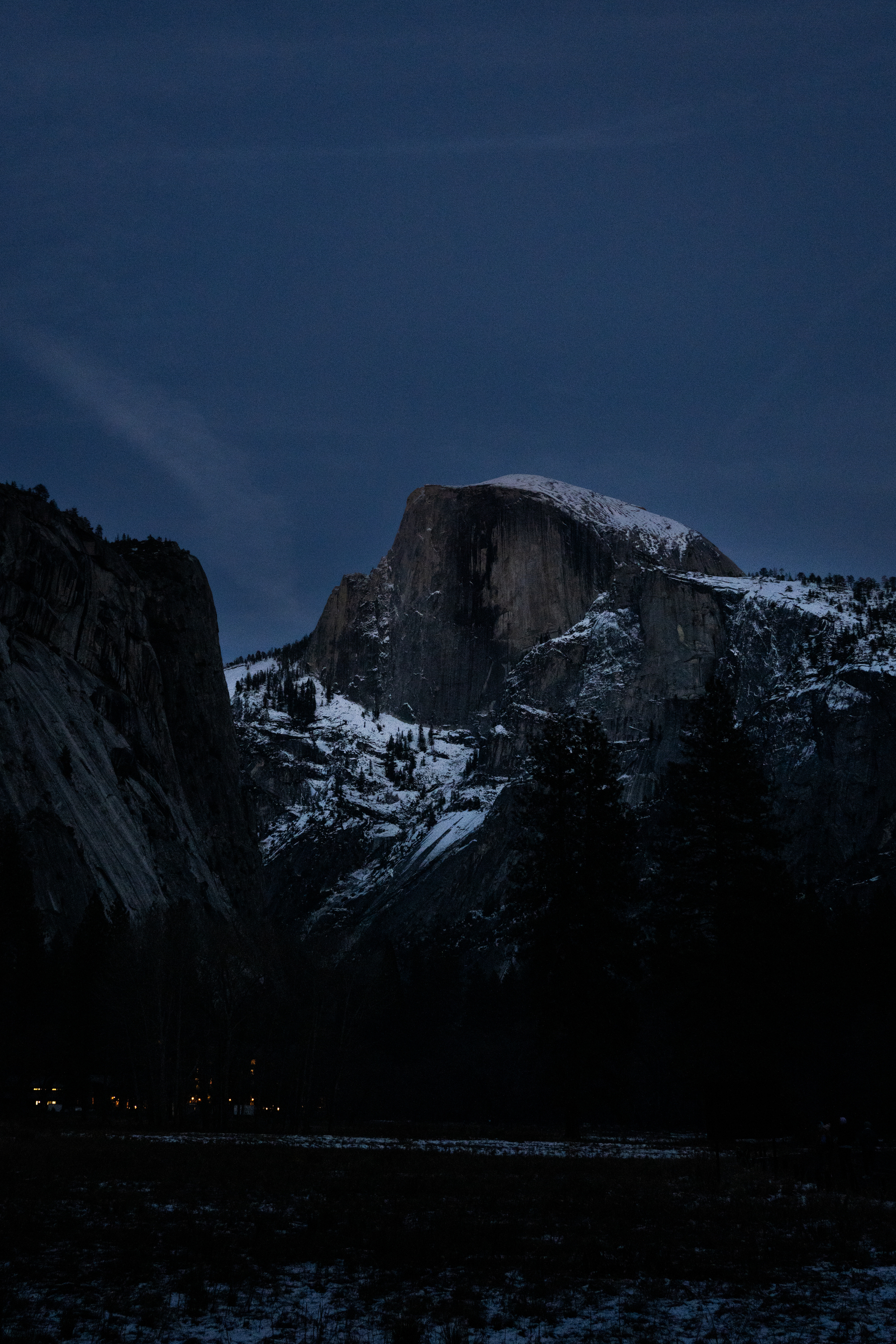 A snow-capped Half dome, in Yosemite National Park, towers over the Yosemite Valley floor, on a dark night, just after sunset.