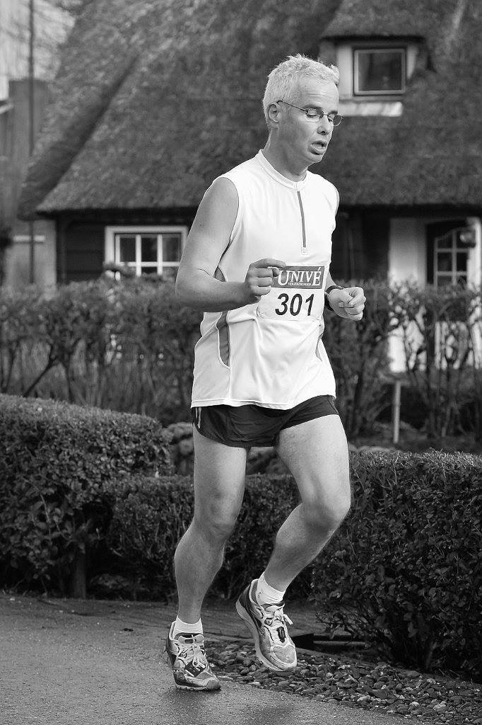 A man is running a race while wearing athletic clothing and a numbered bib in front of a house with a thatched roof.