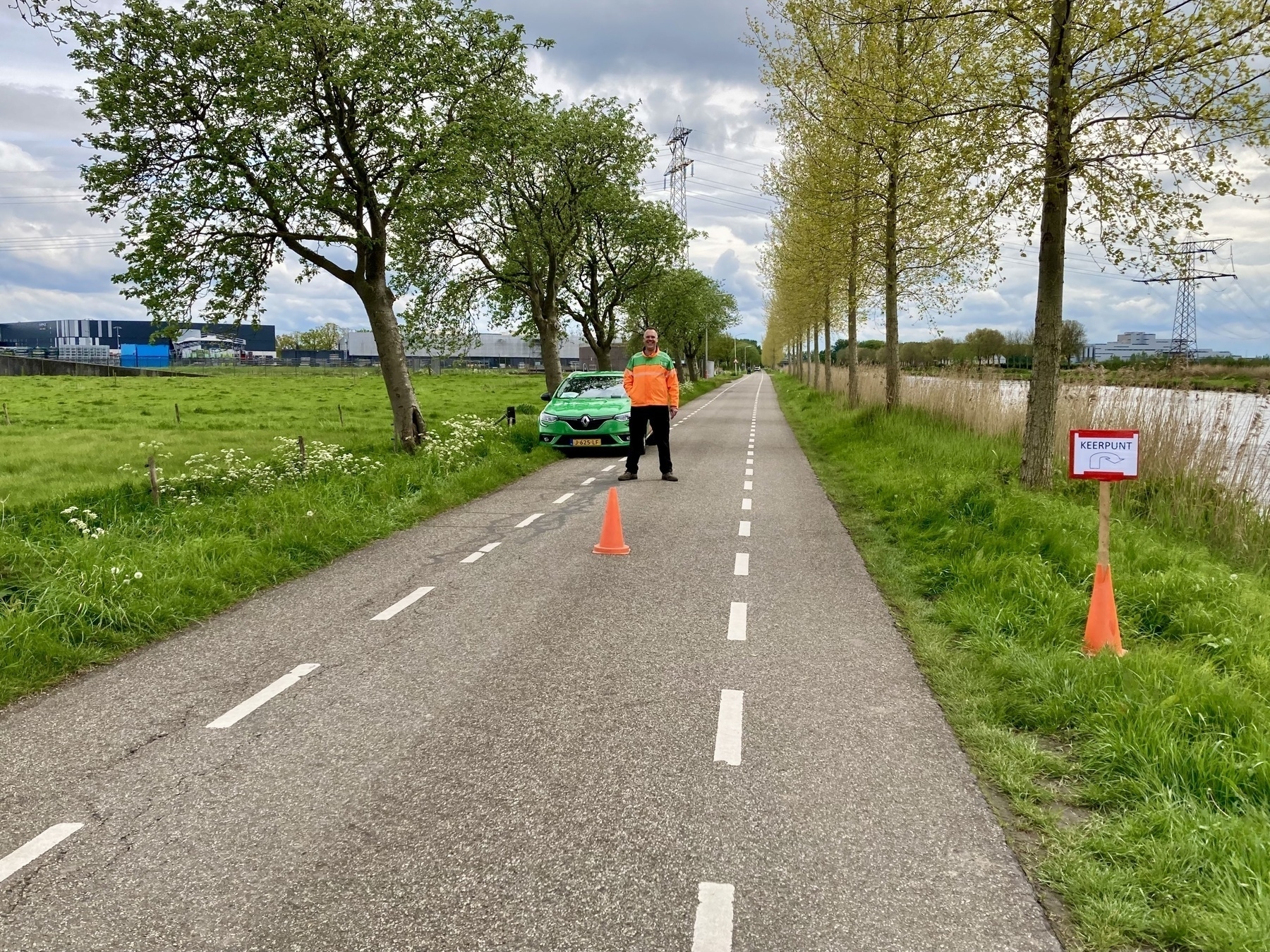 A person wearing a high-visibility vest stands between traffic cones on a small road lined with trees, with a green vehicle parked on the side and a red and white sign that indicates a message for runners to turn here.