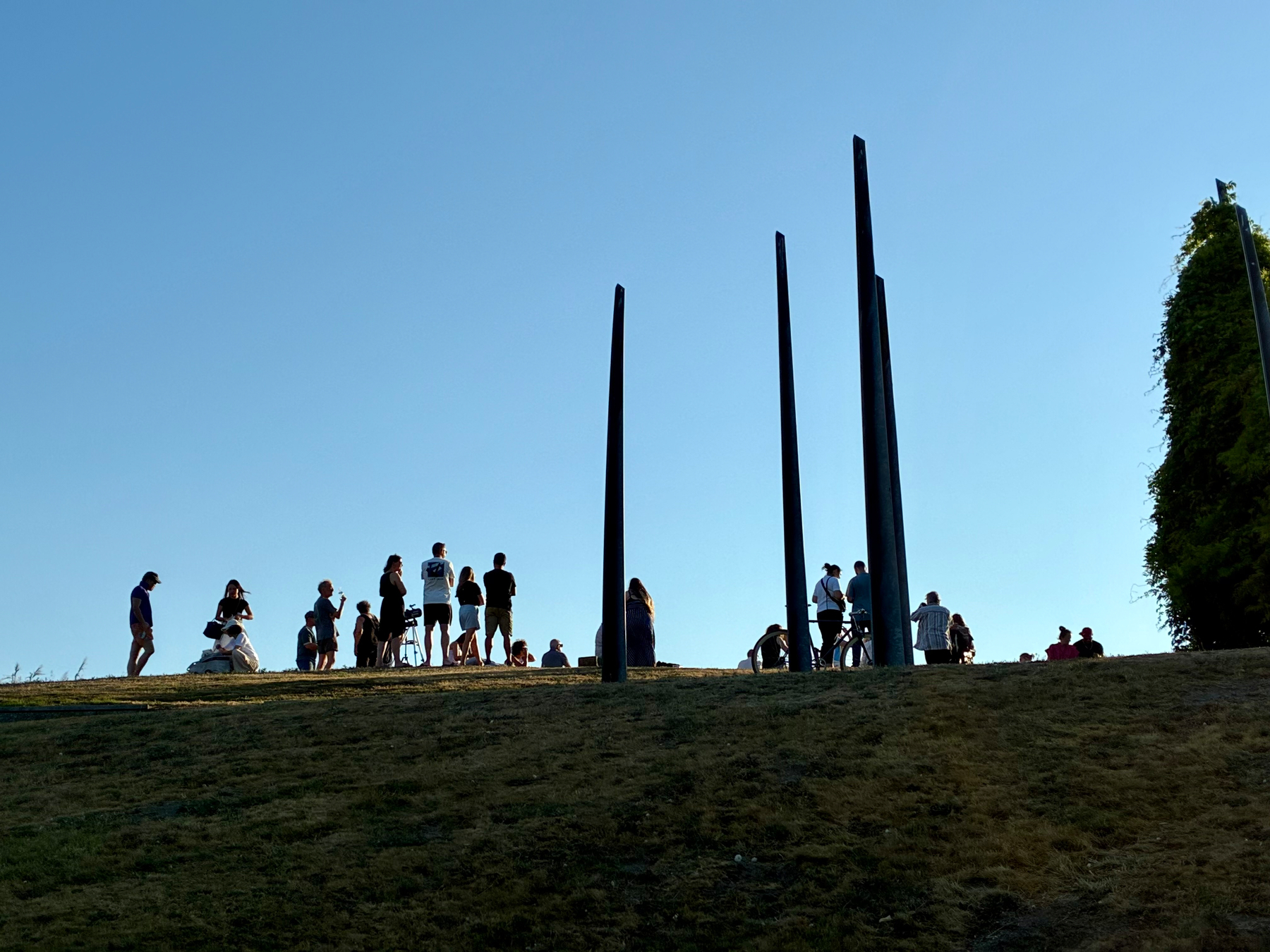 Auto-generated description: People are gathered on a grassy hill near three tall, narrow sculptures against a clear blue sky.