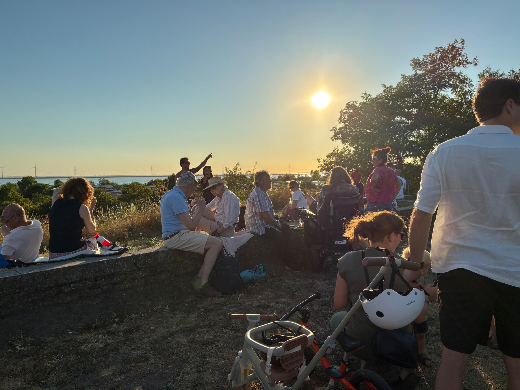Auto-generated description: A group of people are gathered outdoors near a grassy area at sunset, with some sitting on a stone wall and others standing or sitting on the ground.