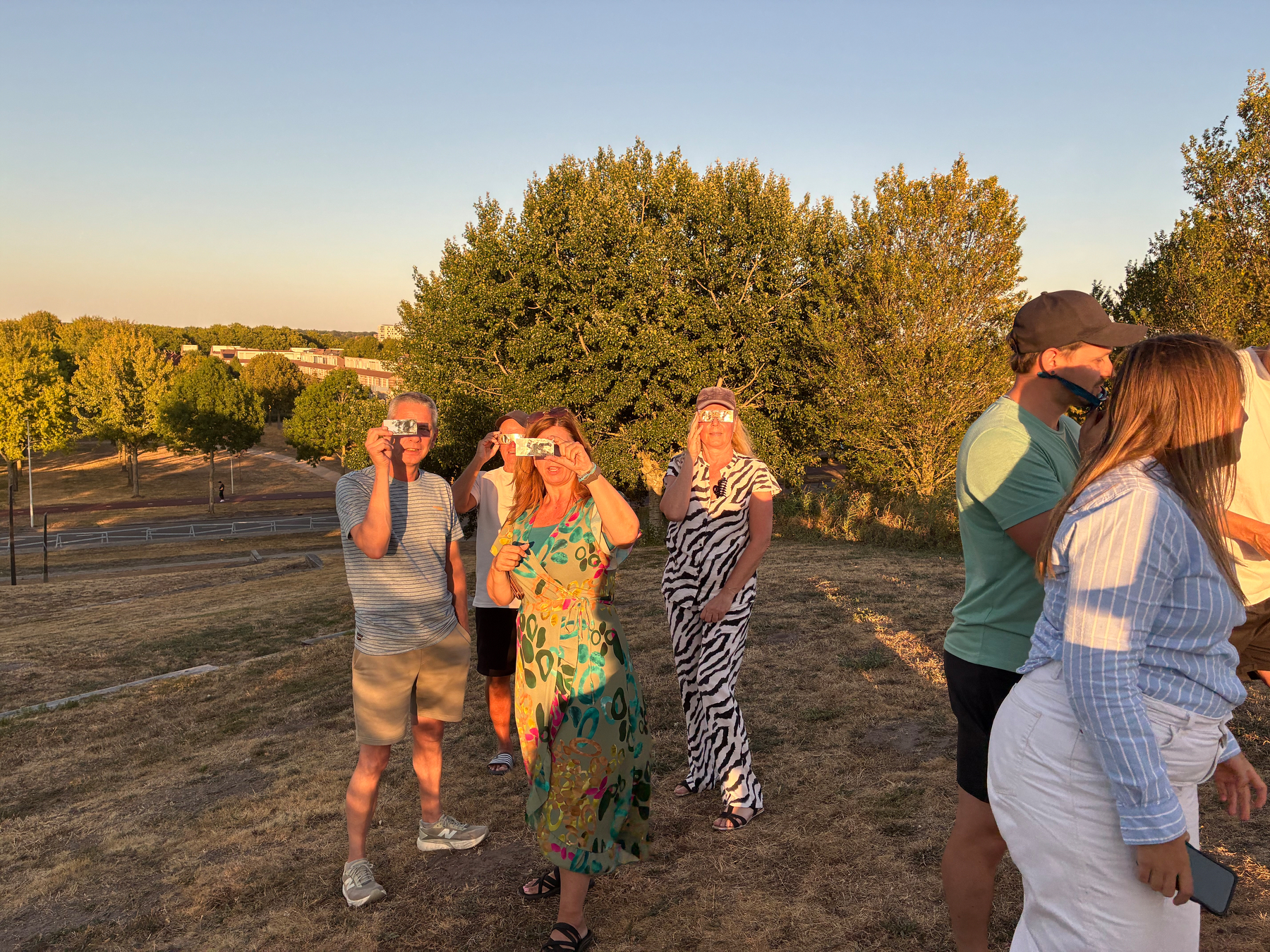 Auto-generated description: A group of people standing on a grassy hill, some taking photos with their phones, with trees and a clear sky in the background.